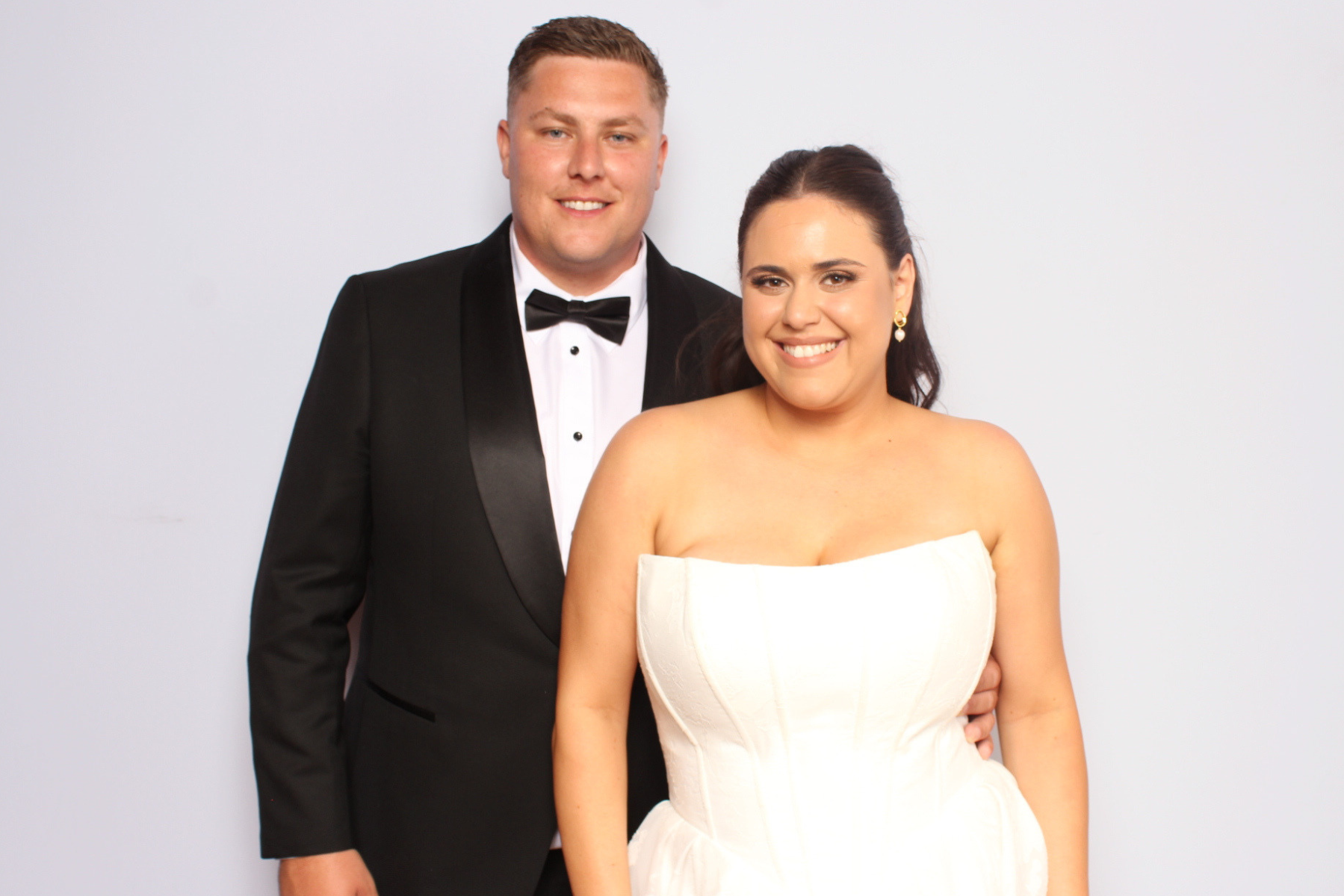 A photo of a couple dressed in formal gowns and tuxedos, standing against a plain white background. The man is wearing a black tuxedo with a white shirt and black bow tie. The woman is wearing a strapless white dress with earrings, and both are smiling at the camera.