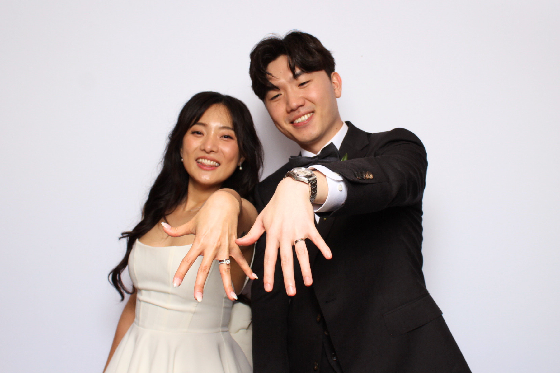 A happy couple dressed in formal wedding attire showing off their rings with both hands extended towards the camera, standing against a plain white background.