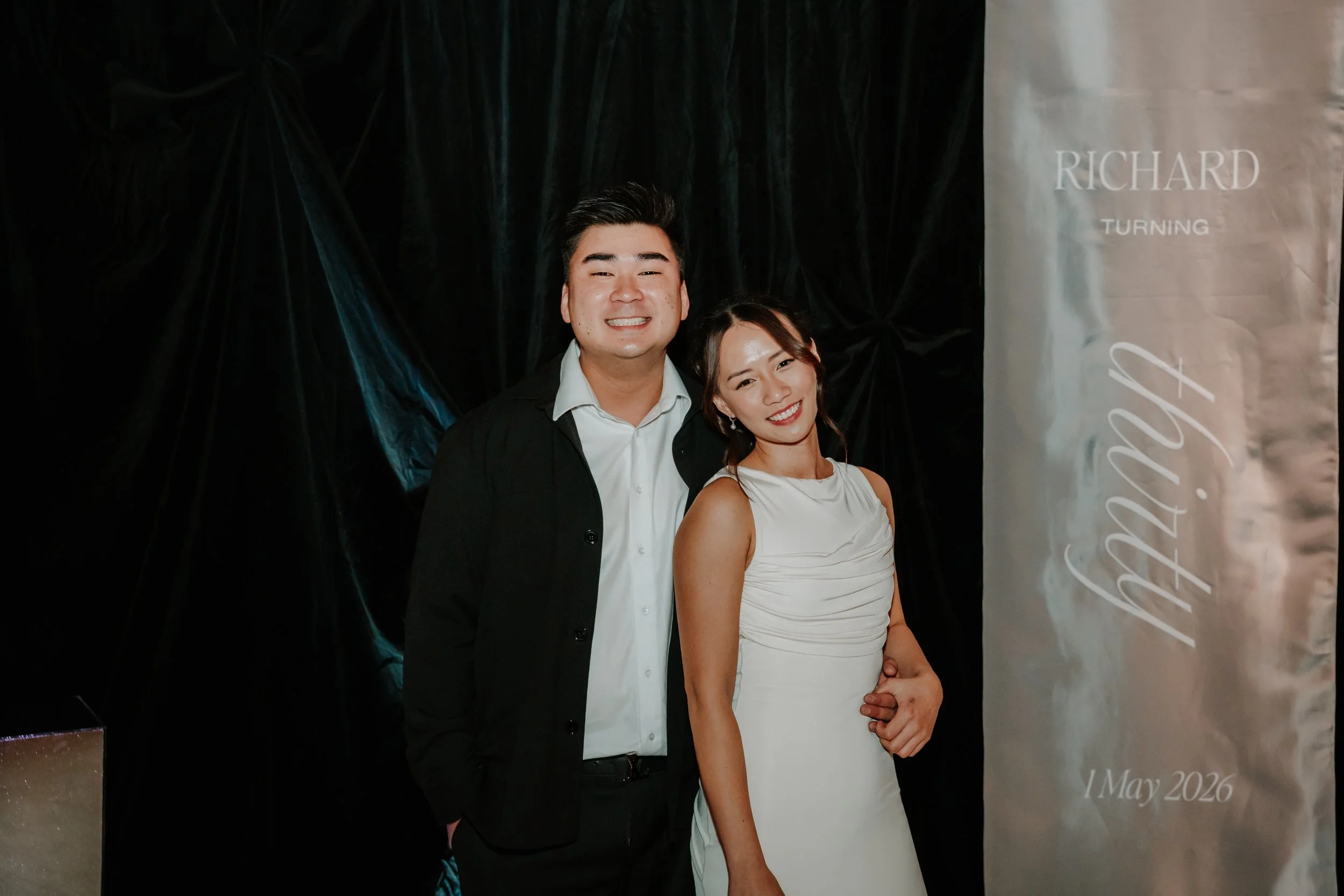 A smiling young man in a black suit and white shirt standing next to a smiling young woman in a white dress, posing at an event with a dark background and a light-colored banner reading "Richard Turning" and "May 2026."