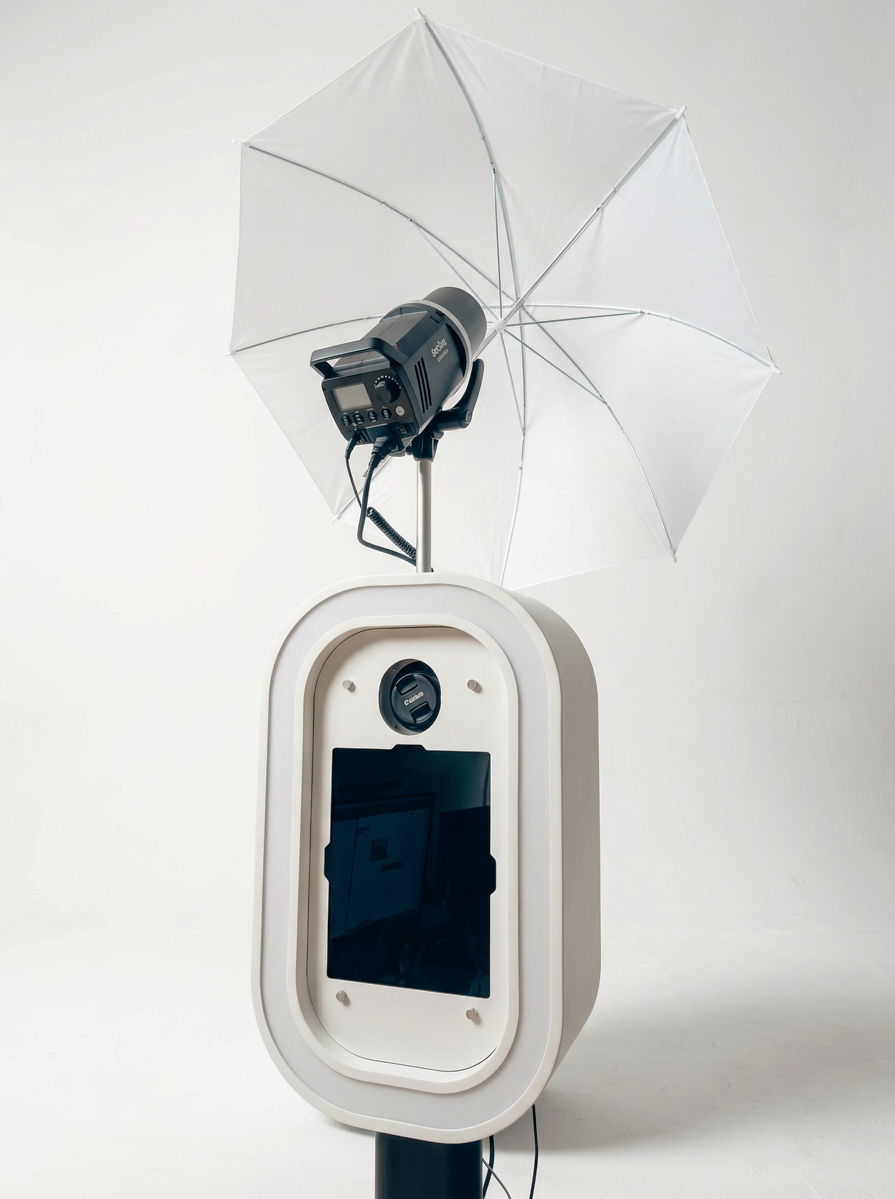 A photobooth with a camera and a screen, topped with a large white umbrella and a portable flash unit.
