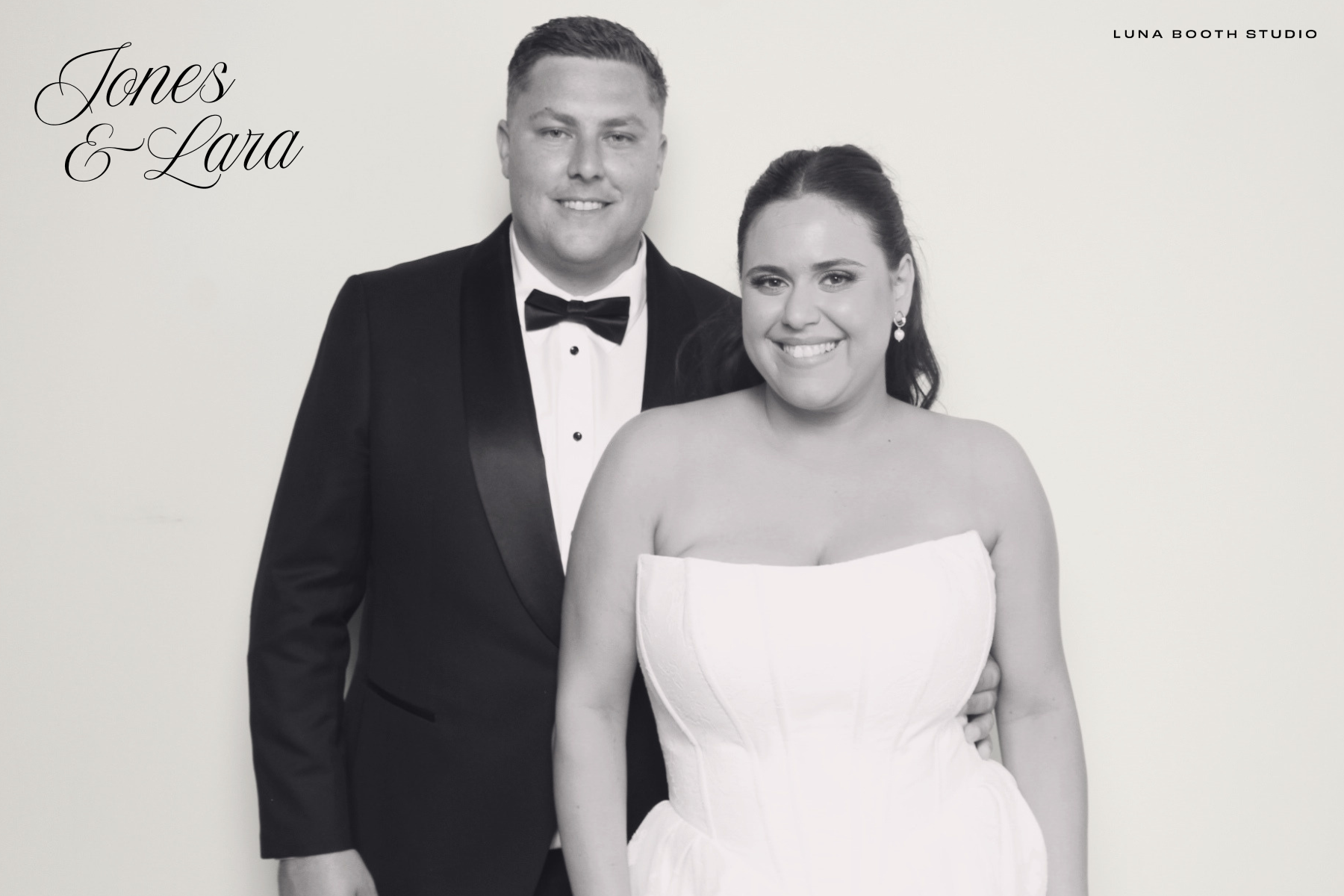 Black and white photo of a smiling man in a tuxedo and a woman in a strapless white dress, both looking at the camera, standing against a plain background with text that says "Jones & Lara" and "Luna Booth Studio."