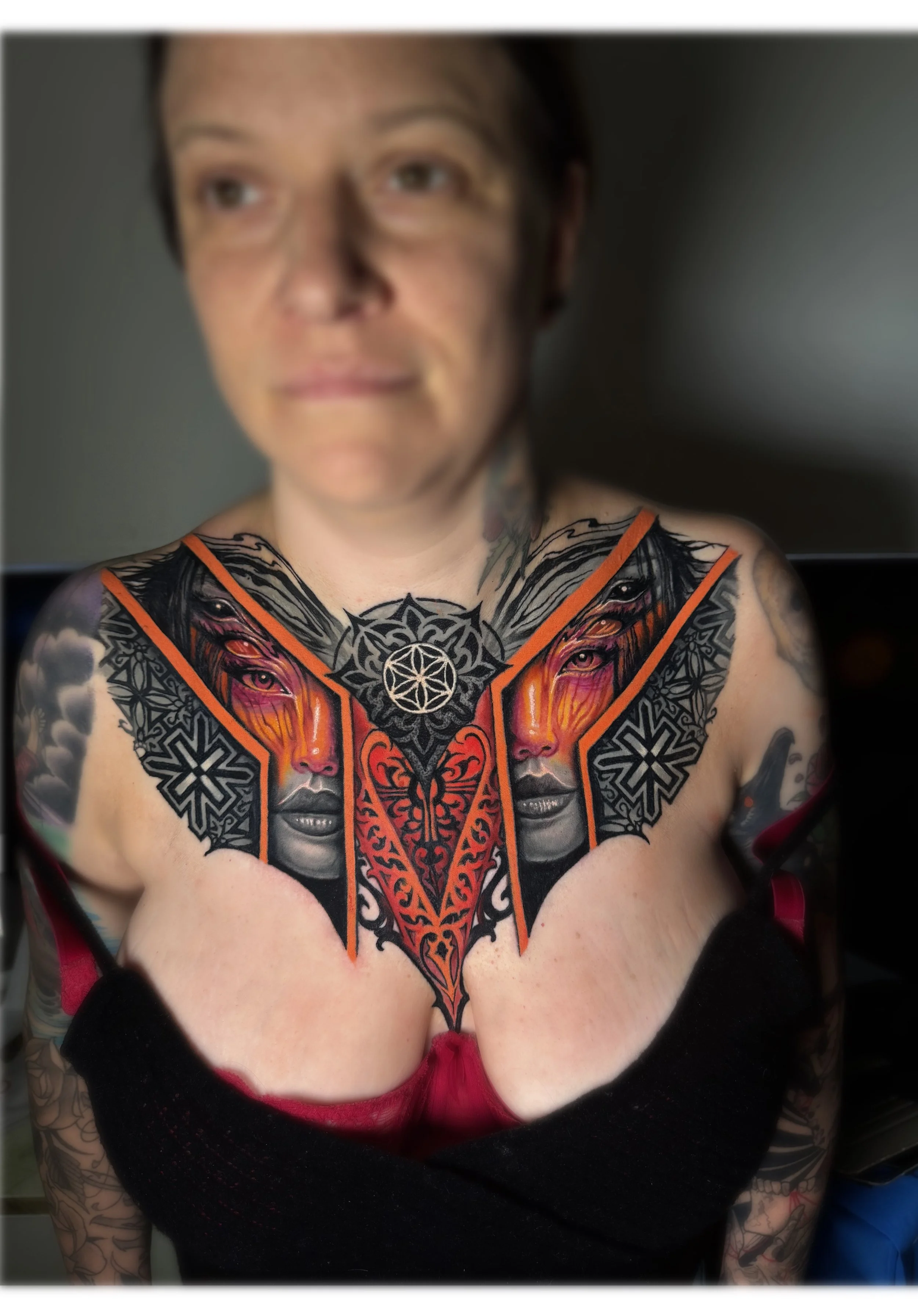 A woman with tattoos on her chest and arms wearing a red and black top, with a blurred background. Daniel Armitage, Calgary Alberta, Surrealist Tattoo Artist, YYC