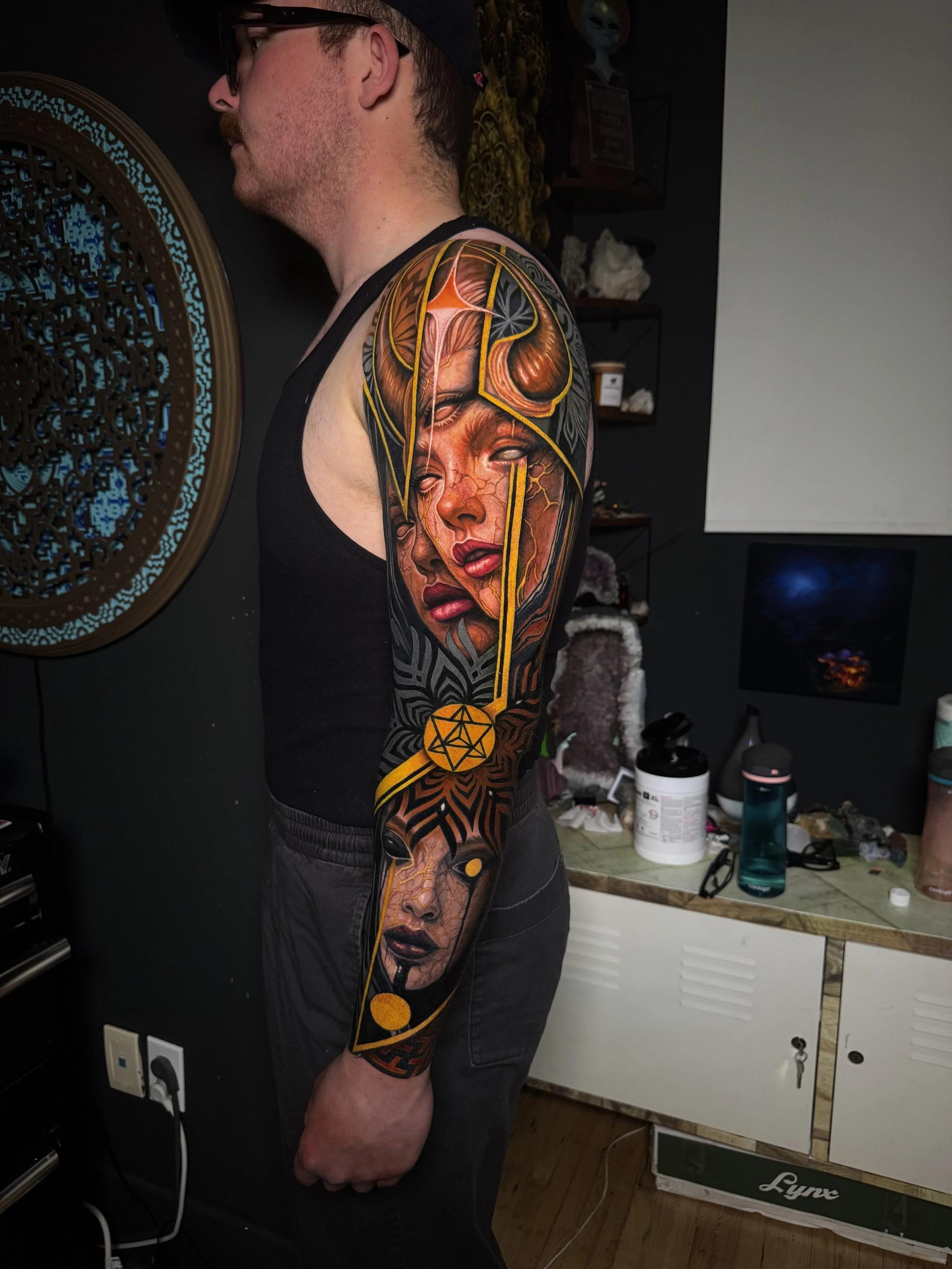 A person with a tattoo sleeve on their arm, wearing a black tank top, standing in a room with shelves, a table, and various objects including bottles, a box, and wall decor. Daniel Armitage, Calgary Alberta, Surrealist Tattoo Artist, YYC