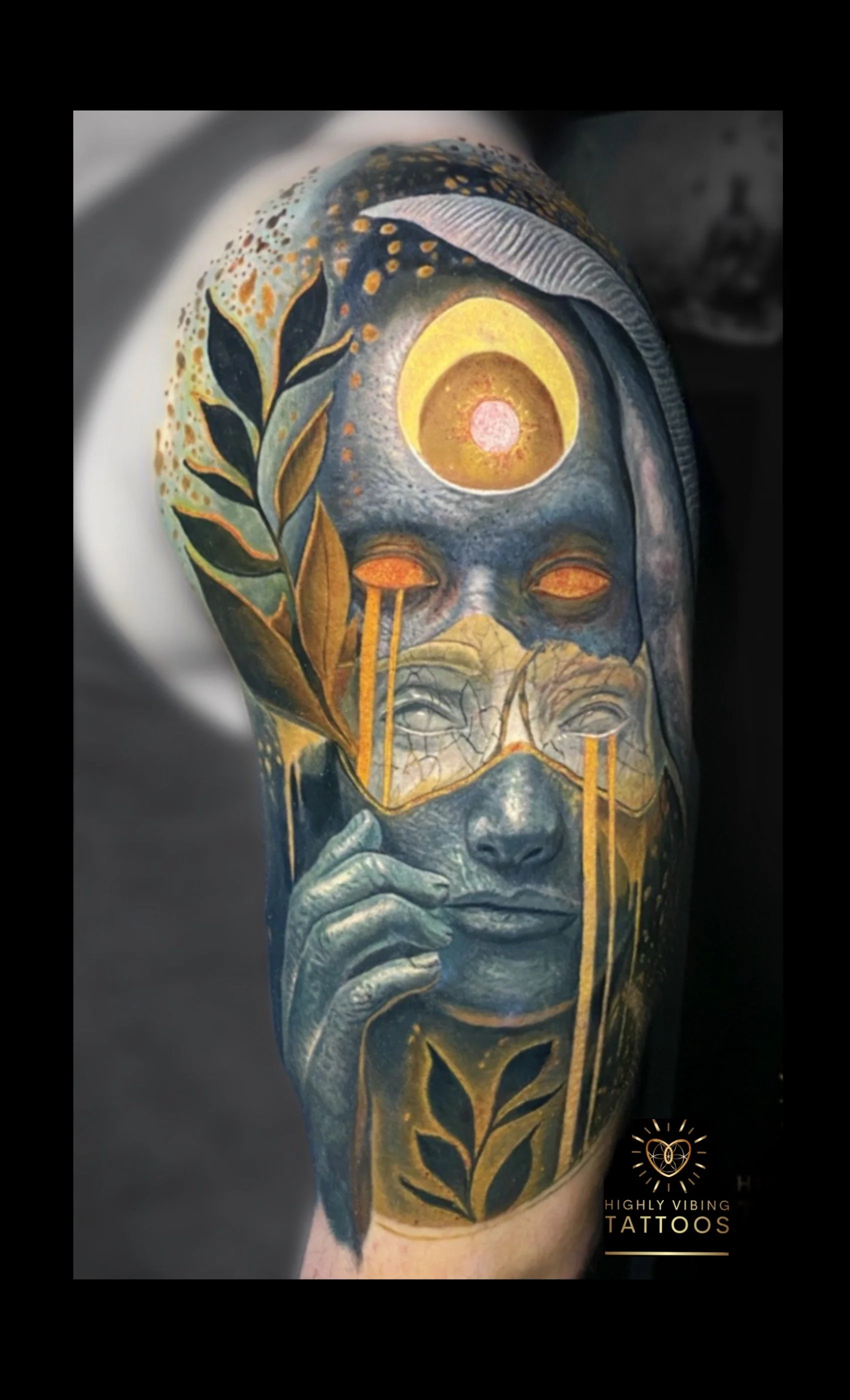 Tattoo of a woman's face with closed eyes, with botanical and celestial elements, including a large eye above her forehead, on a person's arm.
