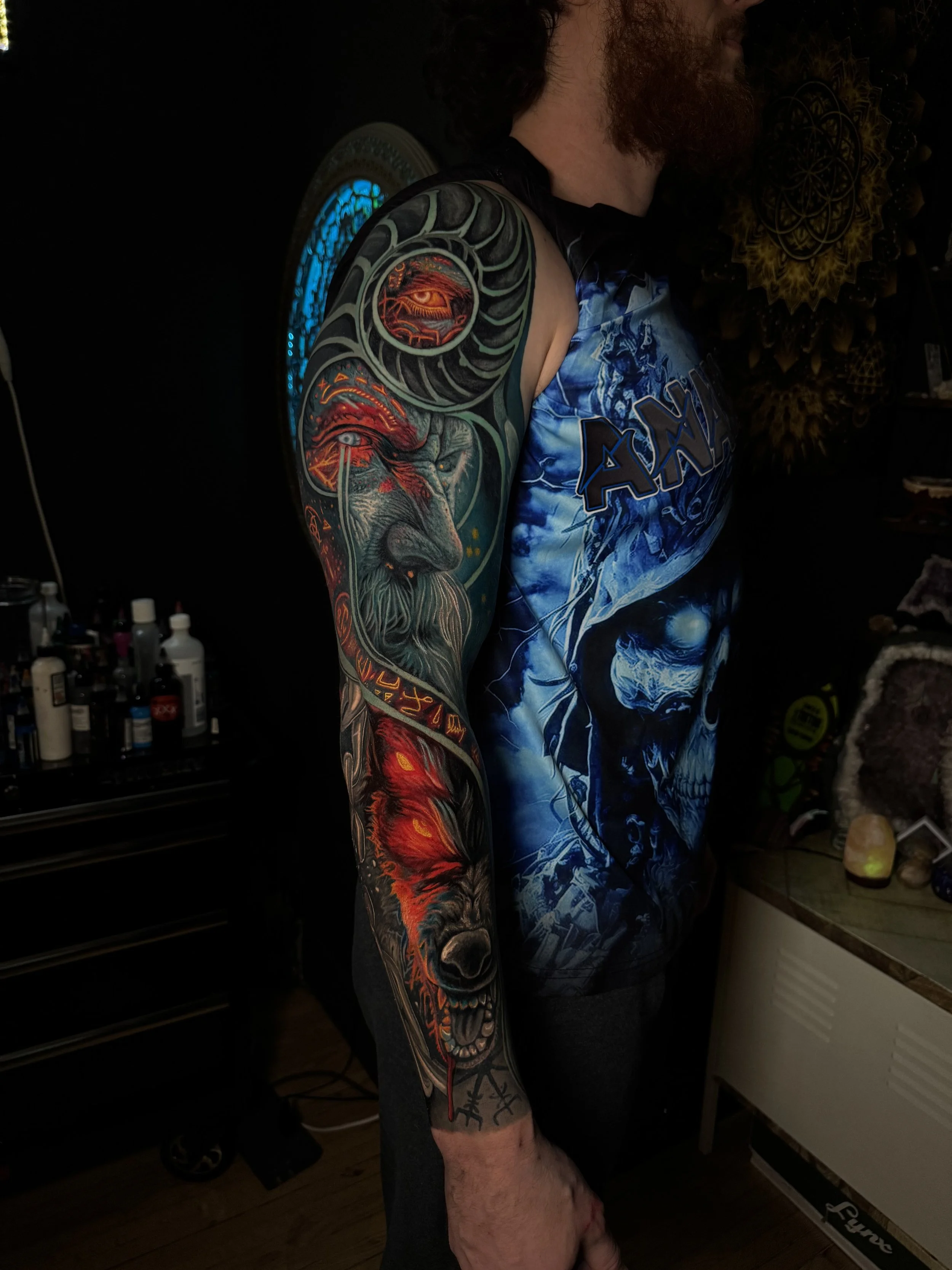 A person with a full arm tattoo featuring a lion's face, an eye, and fire elements. The person is wearing a short-sleeved blue and black shirt and is standing in a room with tattoo supplies and decorative wall art. Daniel Armitage, Calgary Alberta