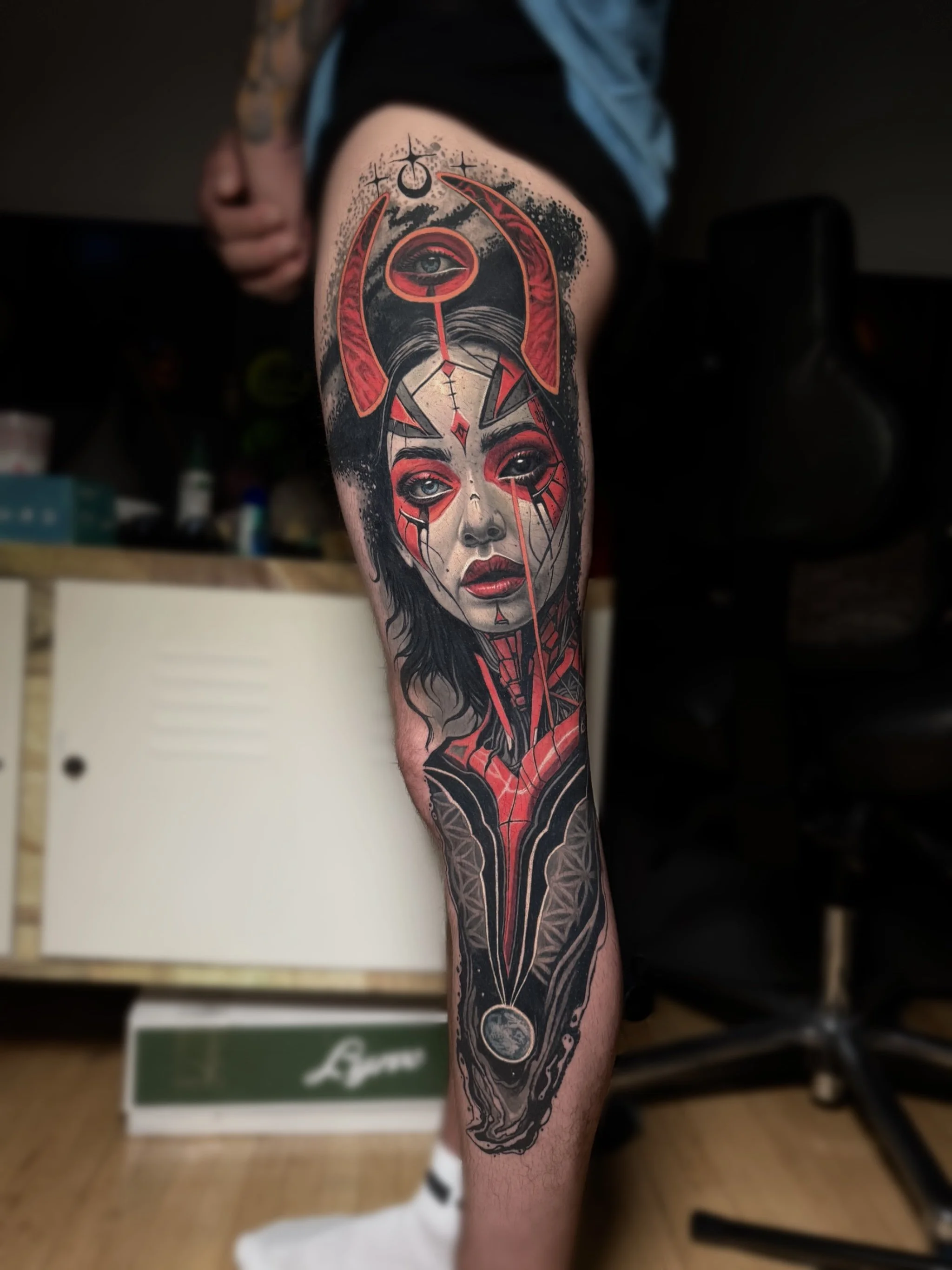 A colorful tattoo on a person's leg features a detailed woman's face with red accents, her hair flowing into abstract black and grey patterns, with lunar and celestial symbols around her. Daniel Armitage, Calgary Alberta, Surrealist Tattoo Artist, YY