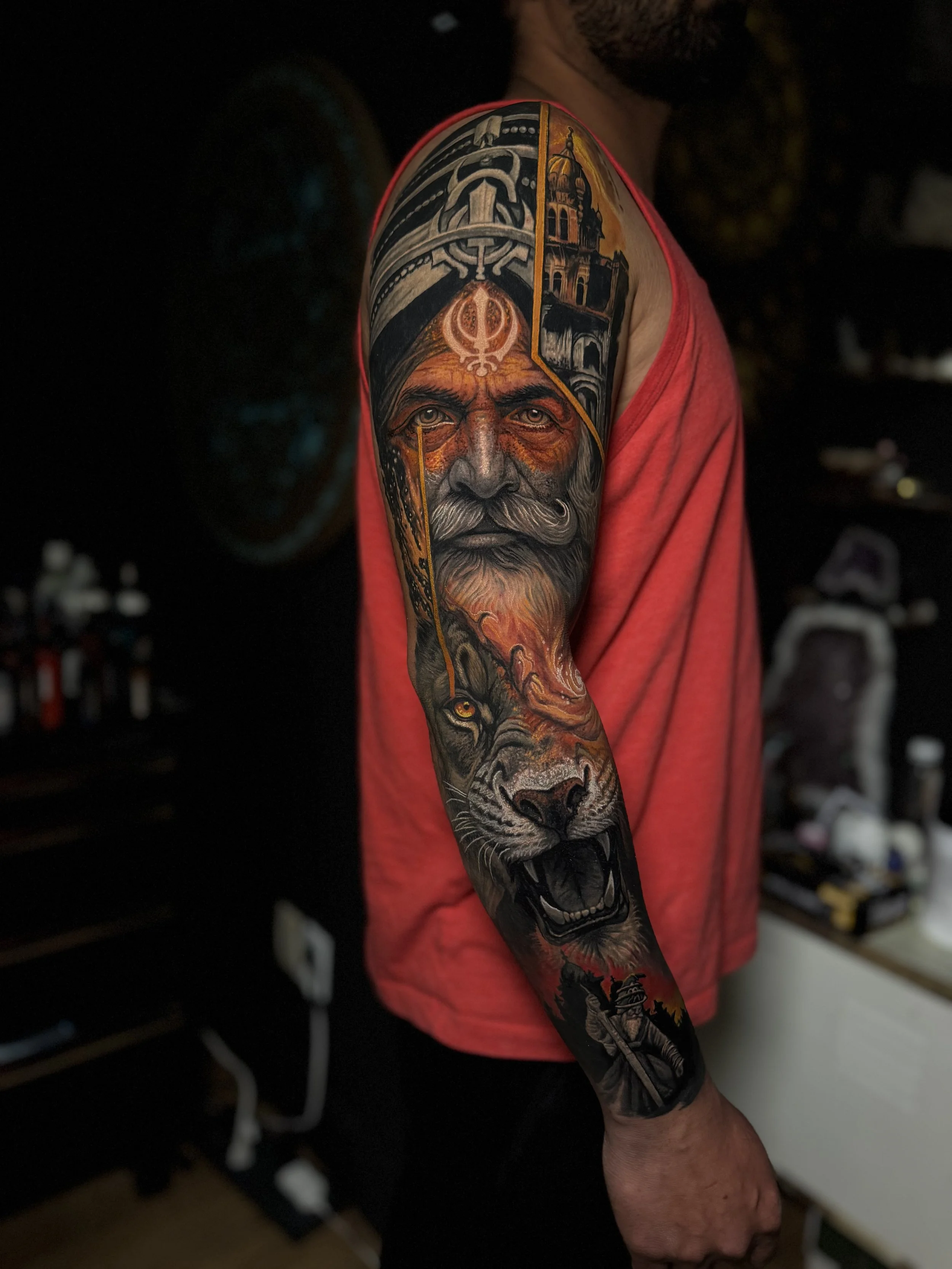 Close-up of a person's arm with a detailed tattoo of a bearded man's face, a wolf, and architectural elements, wearing a red sleeveless shirt. Daniel Armitage, Calgary Alberta, Surrealist Tattoo Artist, YYC