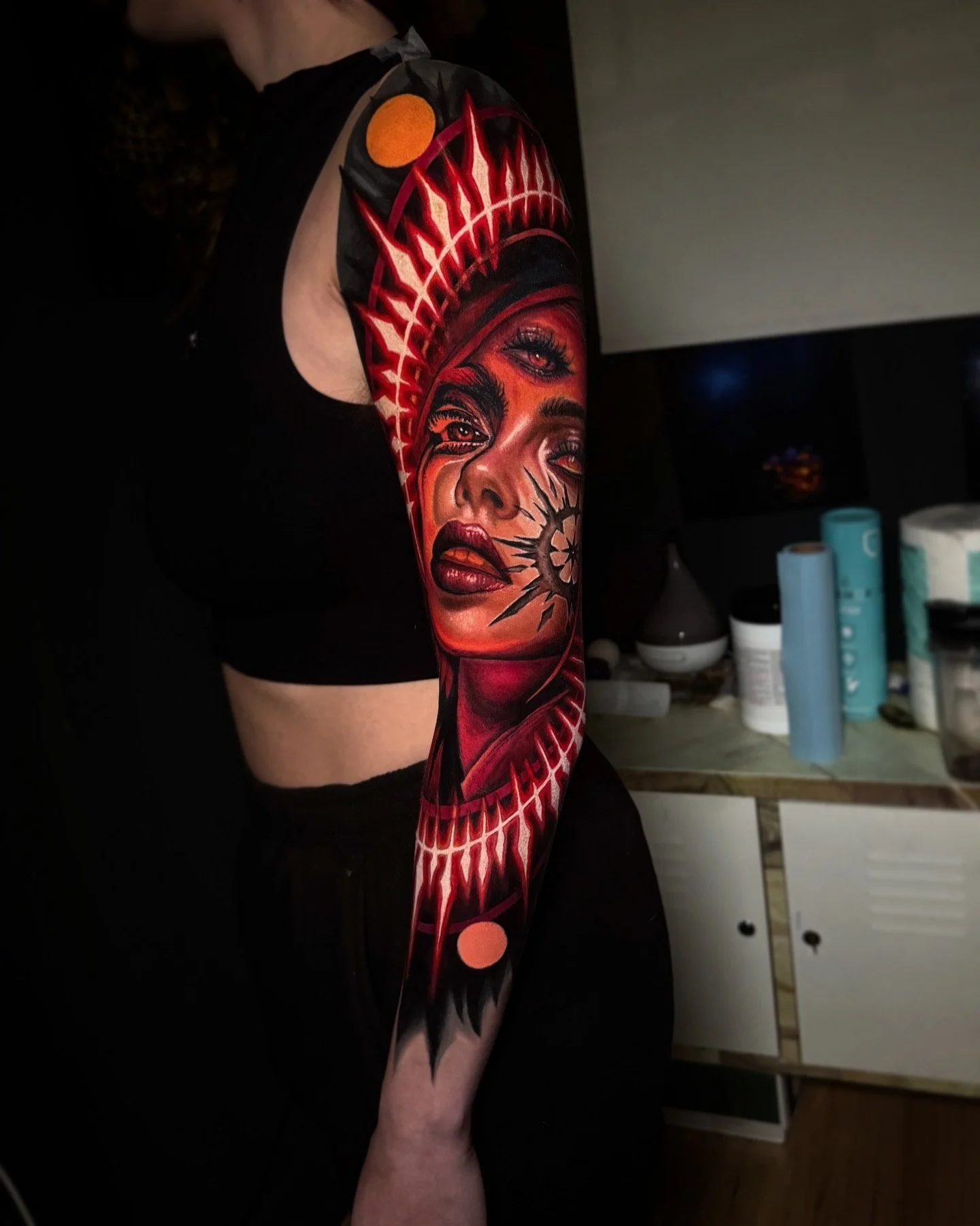 Colorful tattoo of a woman's face on a person's arm, with red, black, and orange details, set in a room with bottles and containers on a counter.