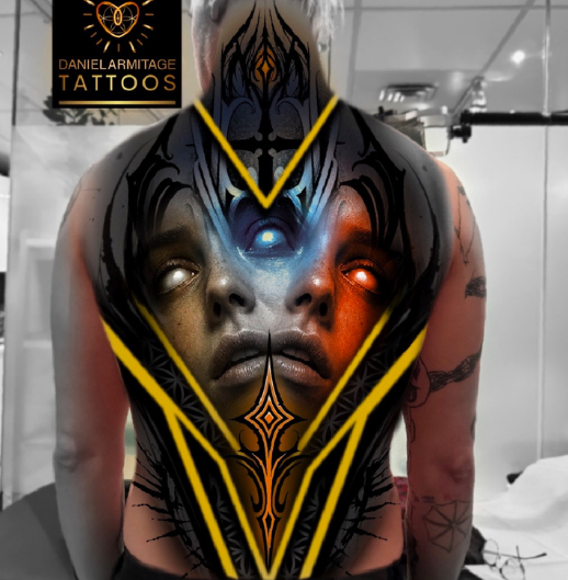 Tattooed person with a black and orange facial tattoo and face paint, posing in a tattoo studio with a DanielsArmitage Tattoos banner in the background. Daniel Armitage, Calgary Alberta, Surrealist Tattoo Artist, YYC