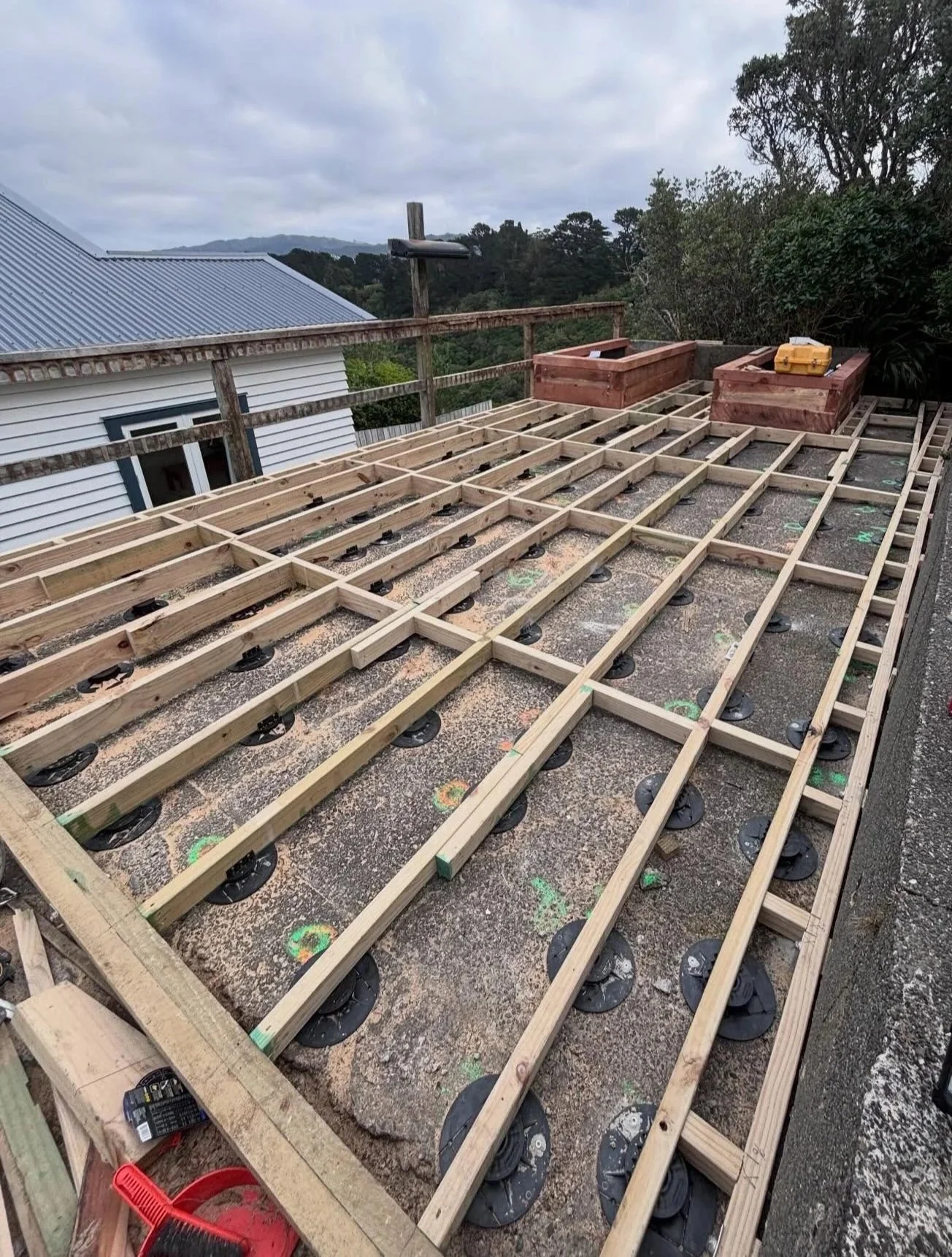 Construction site with wooden framework for a deck or patio, with ventilation discs underneath, on a rooftop or elevated outdoor area, with trees and a cloudy sky in the background.