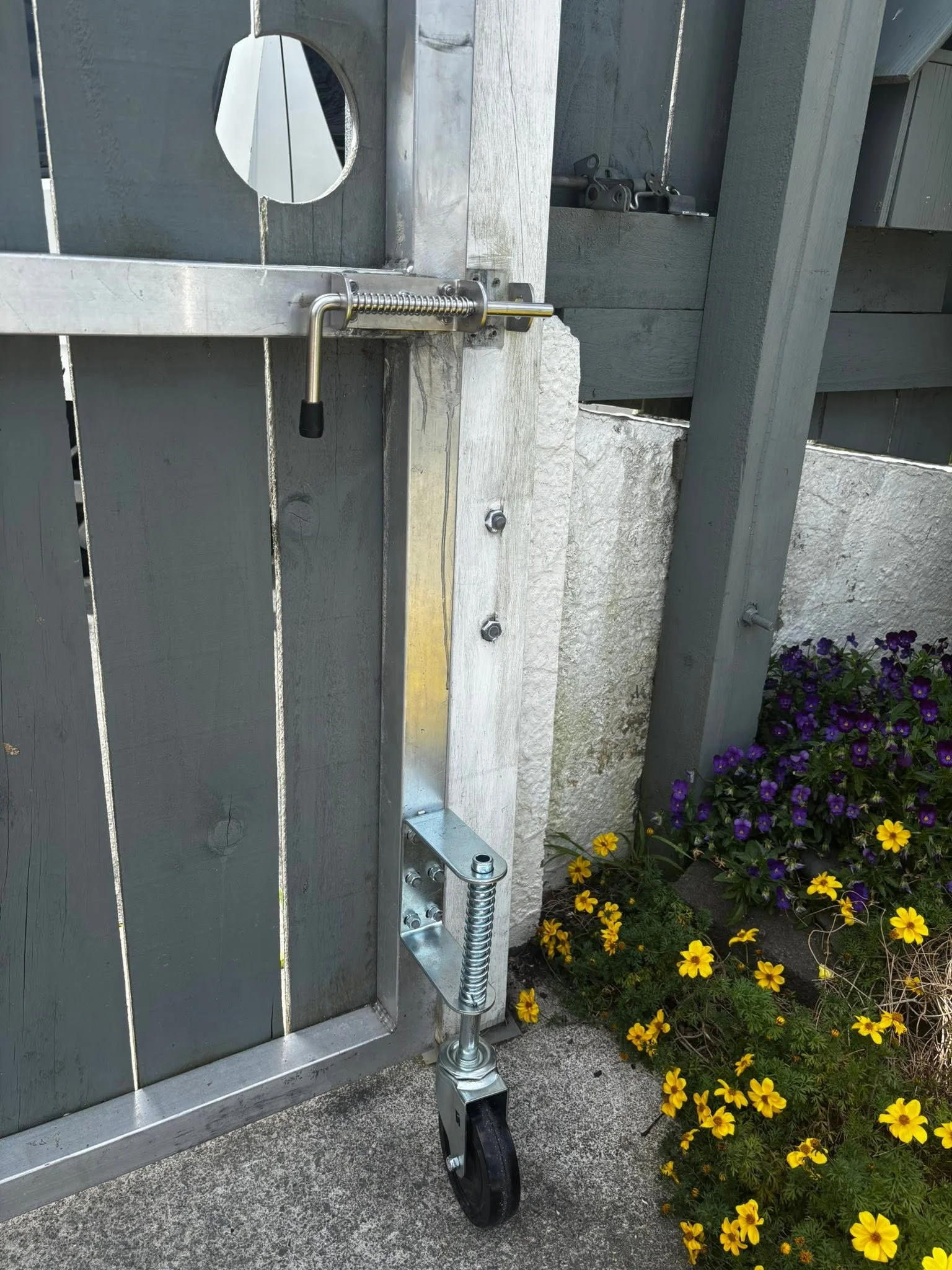 Close-up of a metal gate latch with a spring mechanism and a black wheel, attached to a gray wooden fence, with purple and yellow flowers nearby.