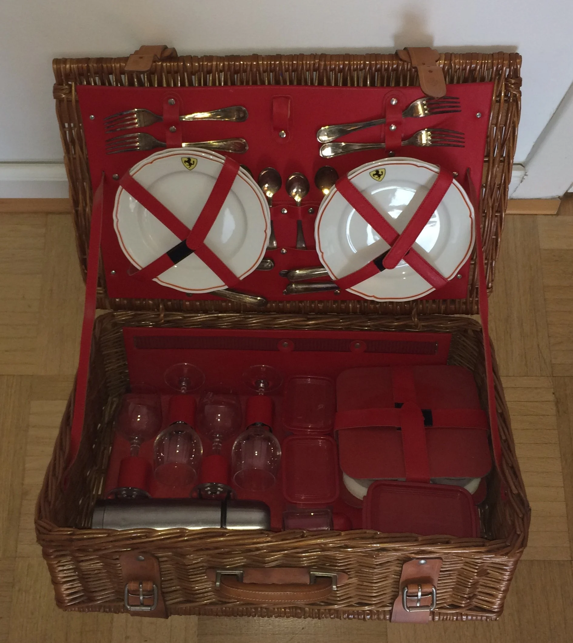 A wicker picnic basket open to reveal neatly arranged silverware, plates, and glasses secured with red straps.
