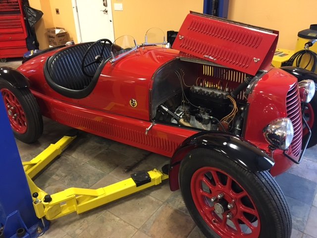A small vintage red race car inside a garage, with its hood open revealing the engine, on a yellow hydraulic lift.