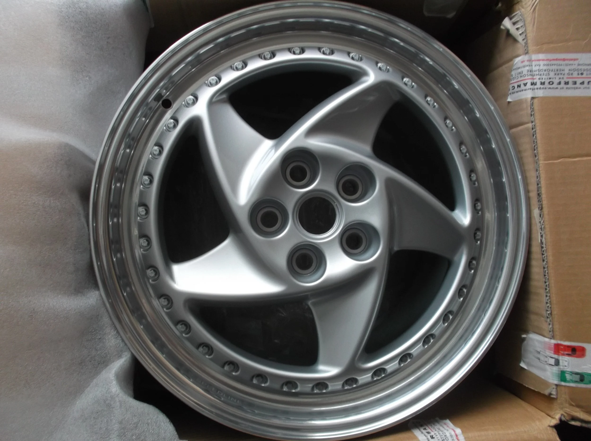 A silver alloy car wheel rim with multiple bolts surrounding the perimeter, placed on a cardboard box.