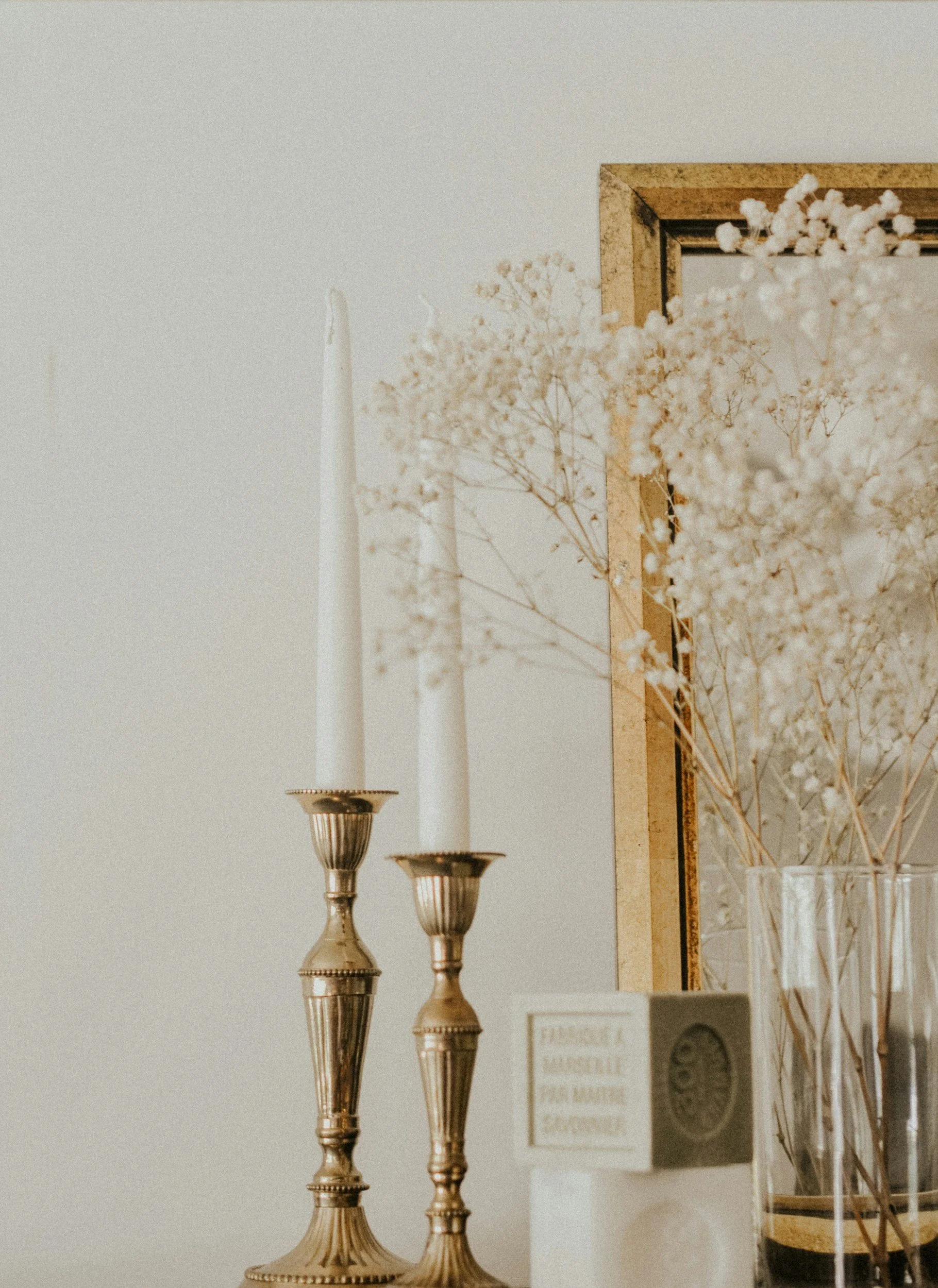 Close-up of three ornate gold candlesticks with tall white candles, and a decorative mirror with a gold frame, alongside a glass vase containing dried white flowers, arranged on a table against a plain wall.