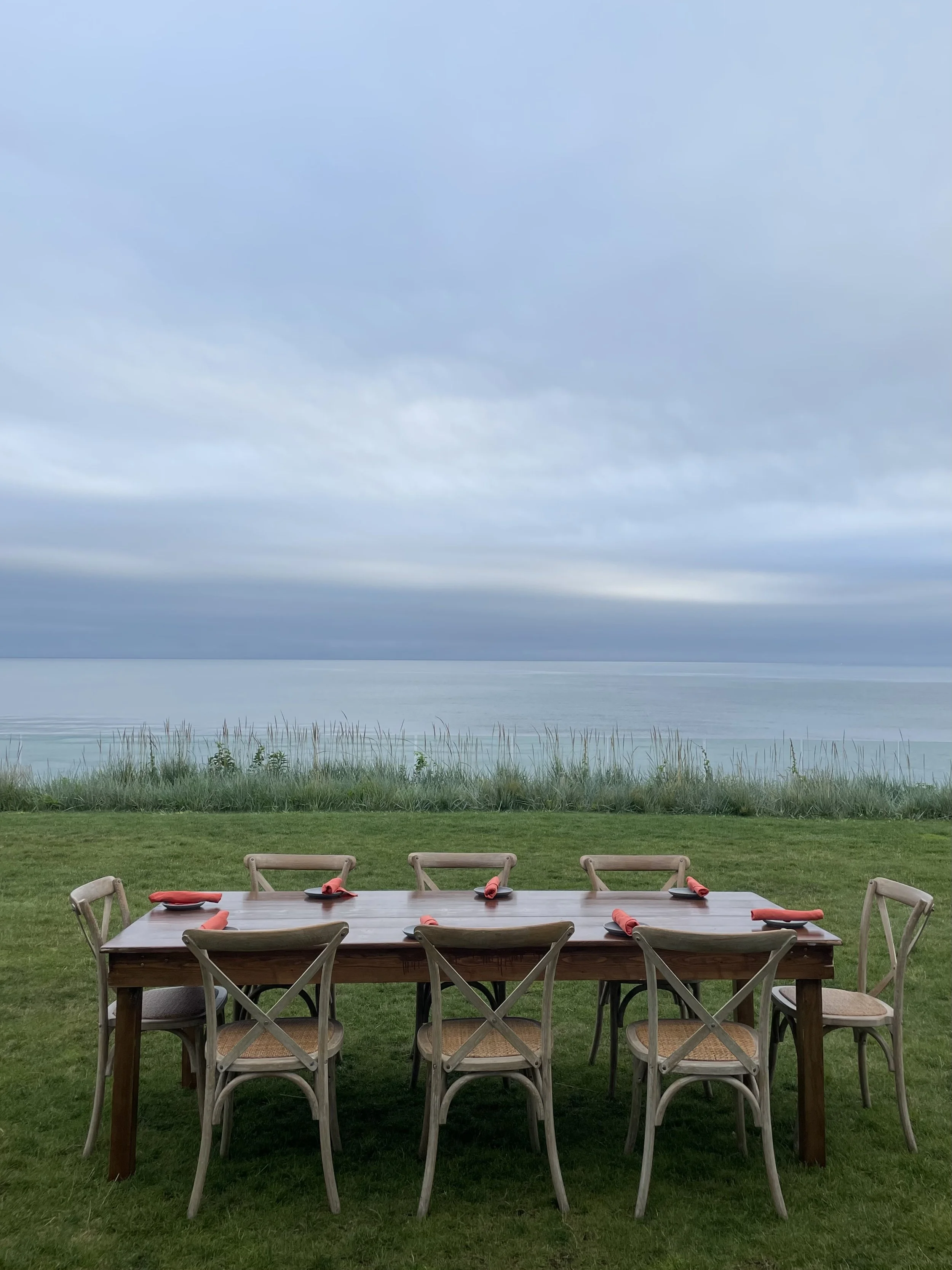 An outdoor dining area with a wooden table and chairs, set with red napkins and black plates, on a grassy lawn with a view of a large body of water, grassy reeds, and cloudy sky in the background.