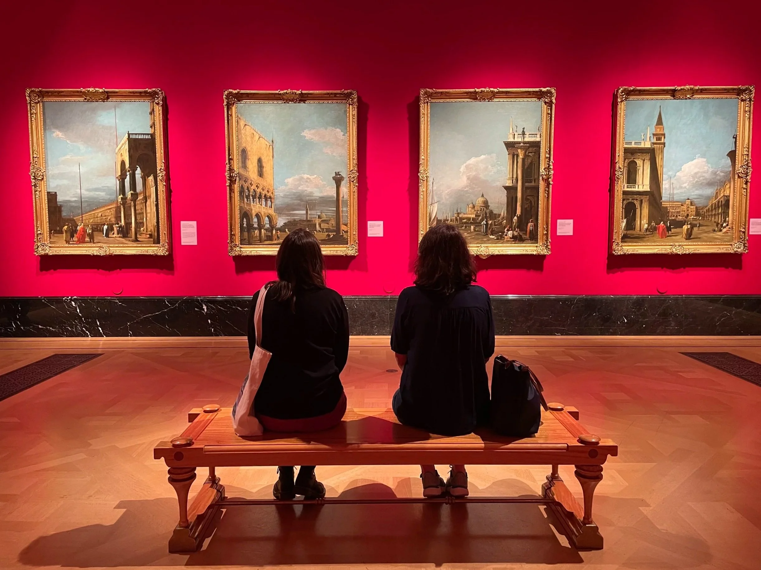Two people sitting on a wooden bench facing four framed landscape paintings in an art gallery with pink walls.