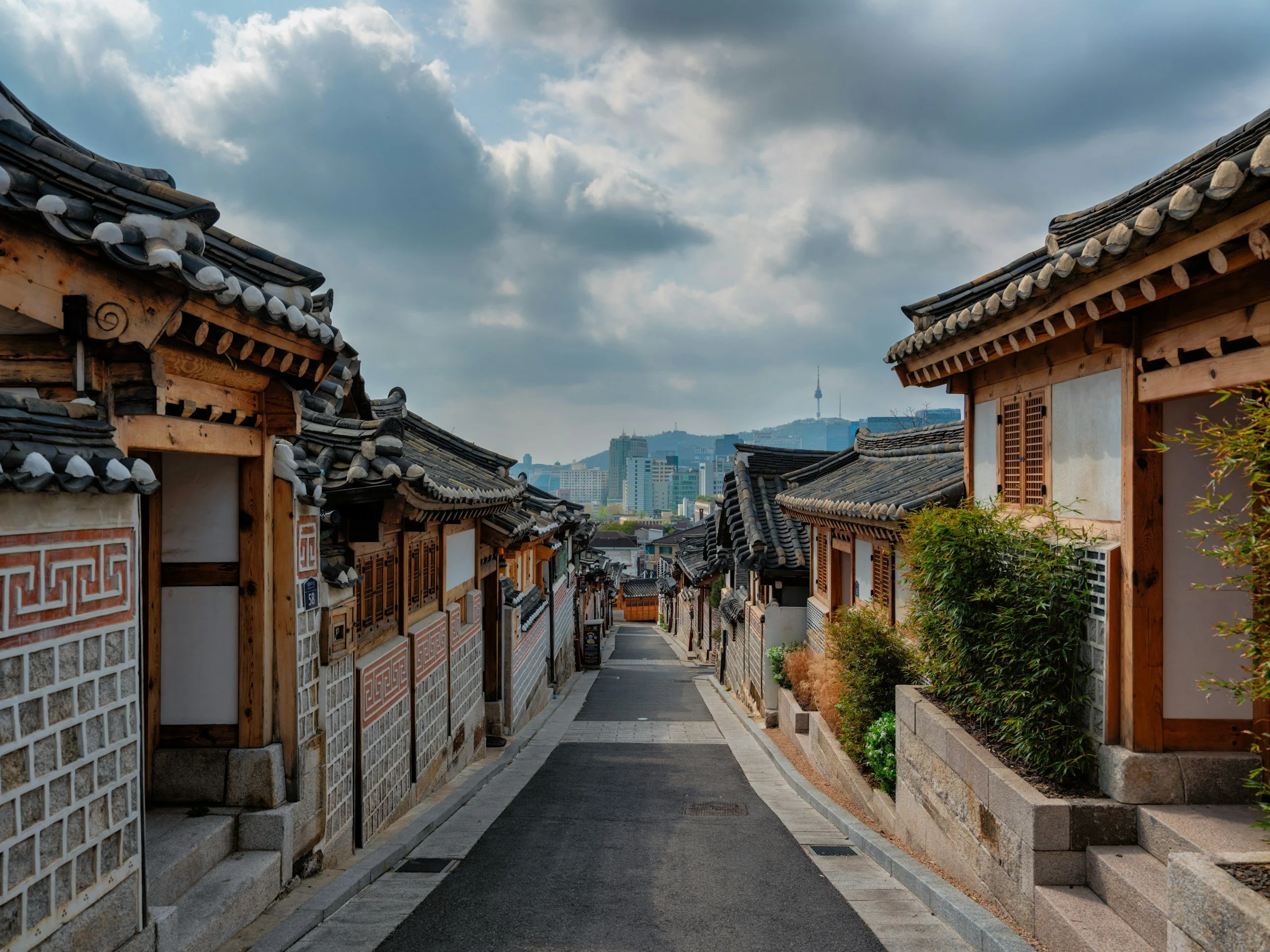 Traditional Korean hanok houses with tiled roofs and wooden beams along a narrow street, with a modern cityscape and N Seoul Tower in the background under cloudy skies.