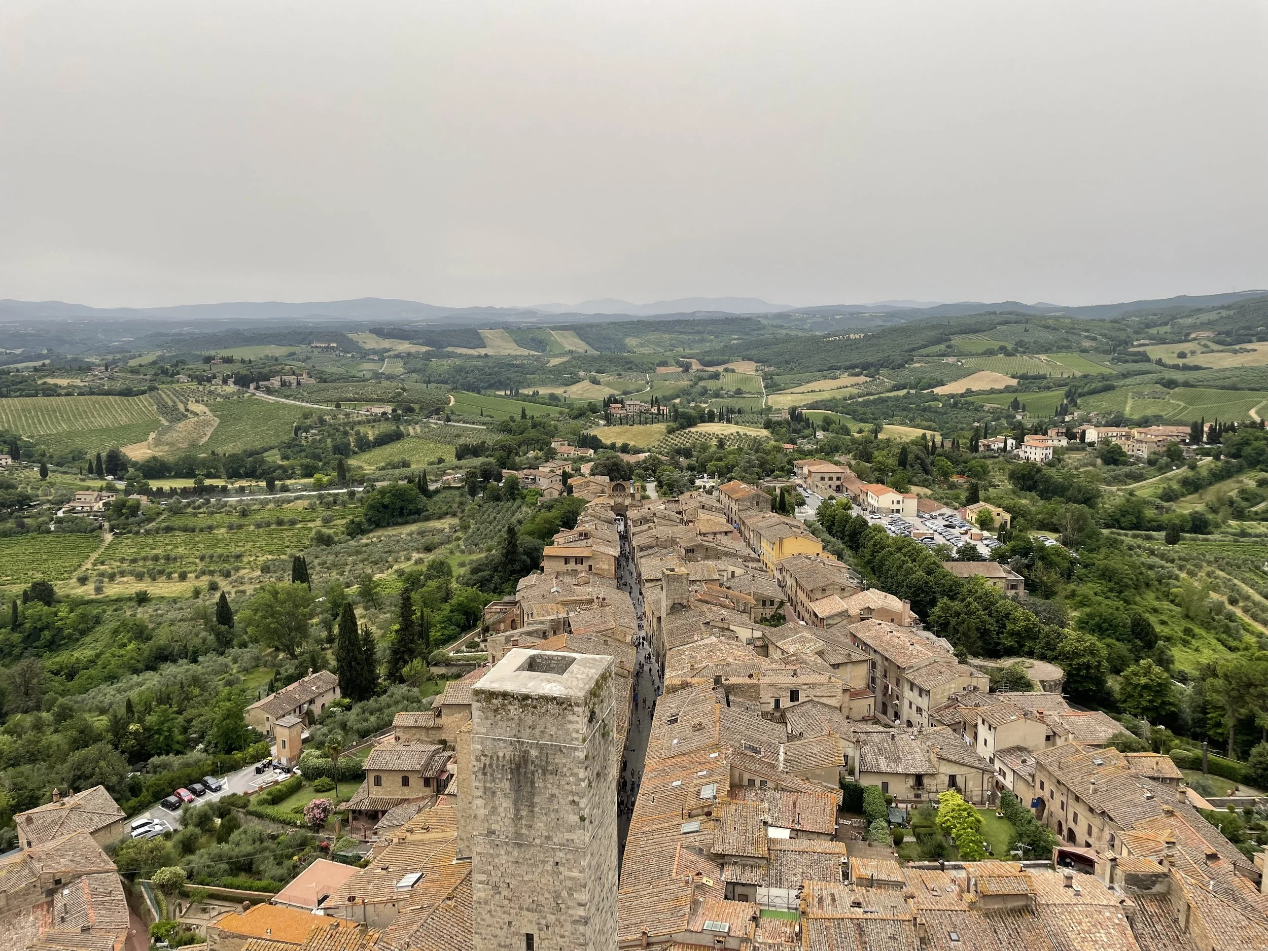 A scenic view of rolling countryside with green fields, vineyards, and distant hills, with an ancient stone tower in the foreground and a small village below.