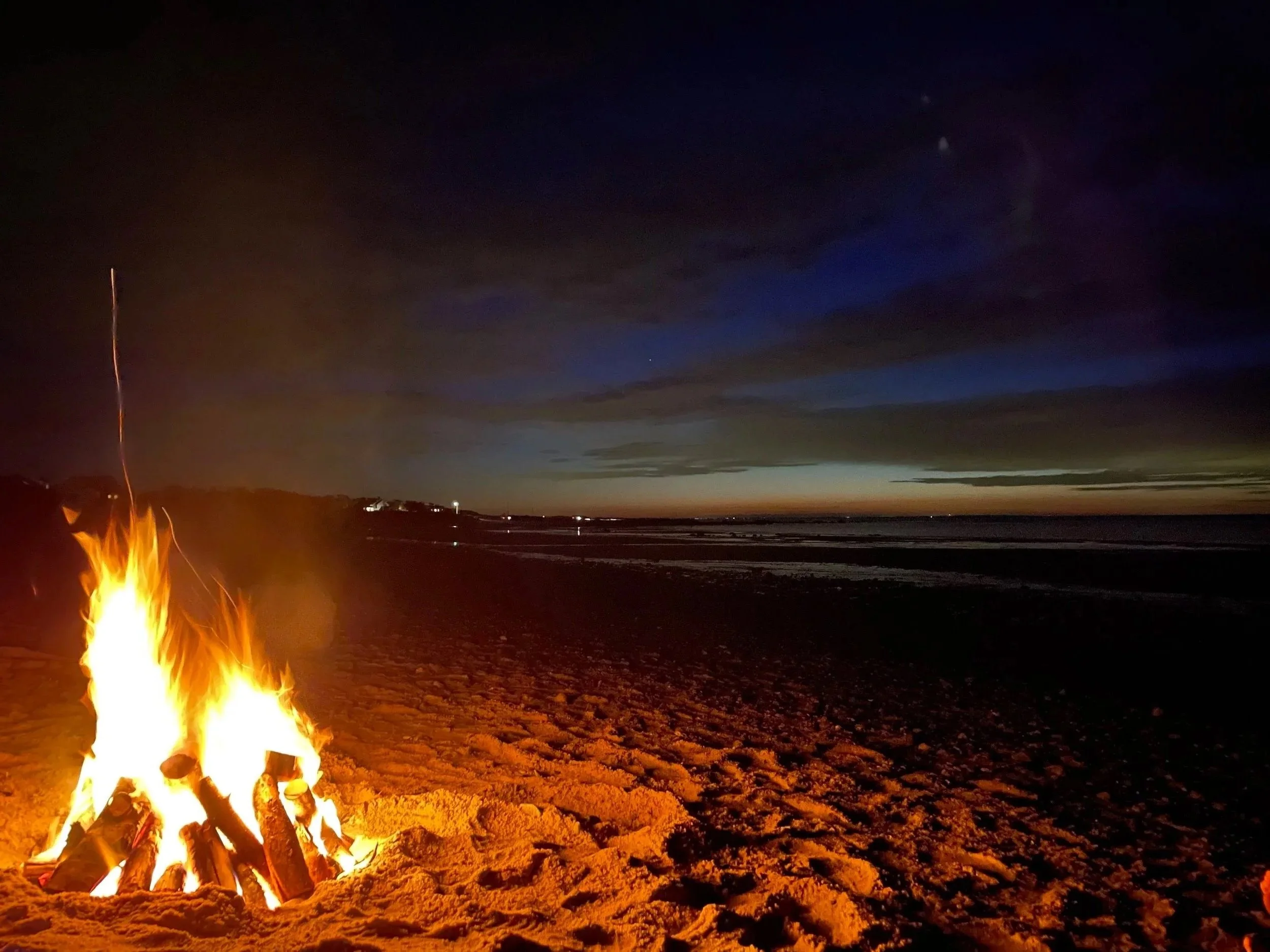 A campfire on a sandy beach at dusk with the ocean and a dark sky with clouds and stars in the background.