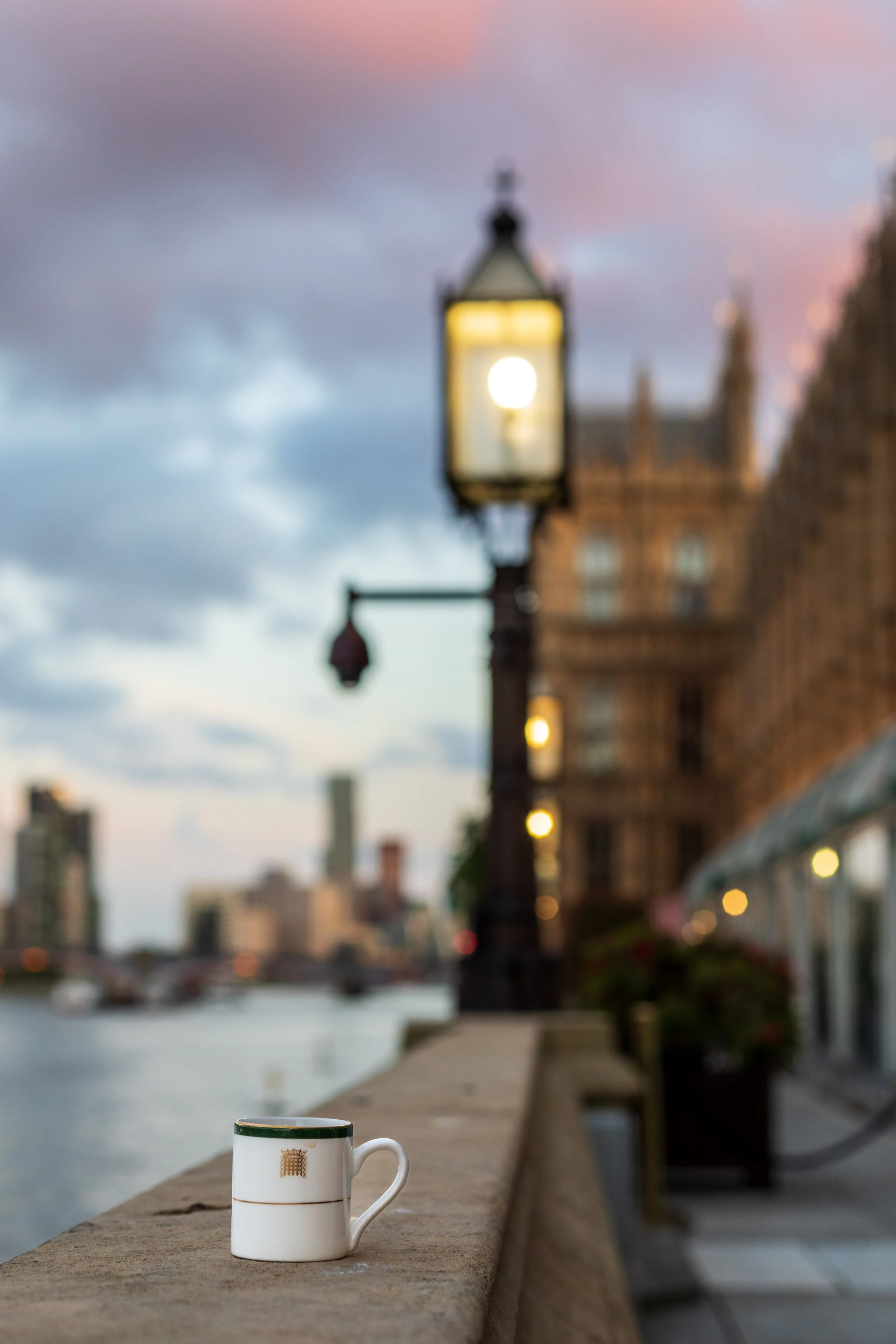 A white coffee mug with gold accents and a coat of arms sits on a concrete ledge next to a river, with blurred buildings and a cloudy sky in the background during dusk.
