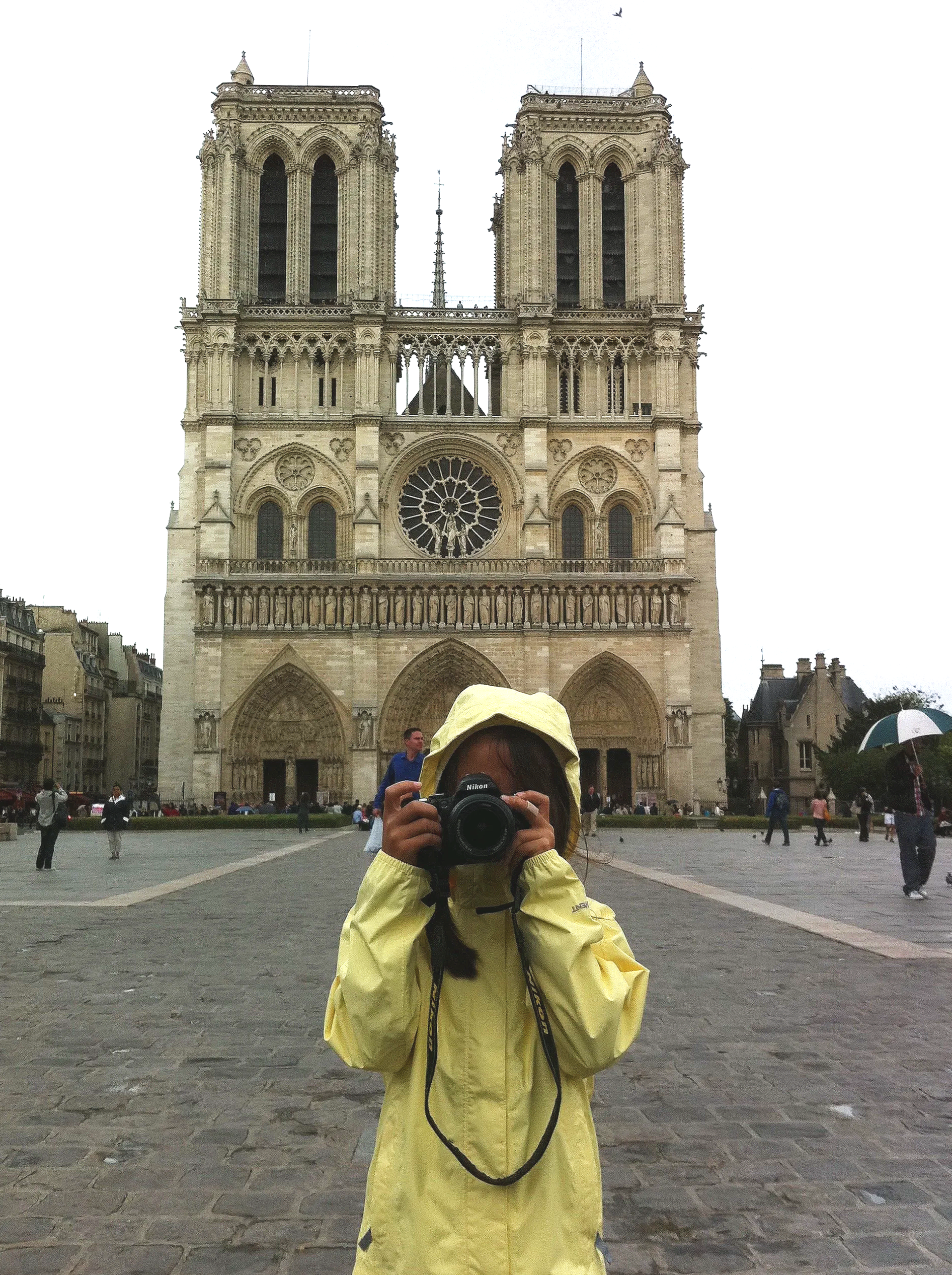 A young girl wearing a yellow rain jacket and hat holding a Nikon camera, taking a photo in front of Notre Dame Cathedral in Paris, France. The cathedral's facade with twin towers and rose window is visible in the background, with people walking and standing around on a cloudy day.