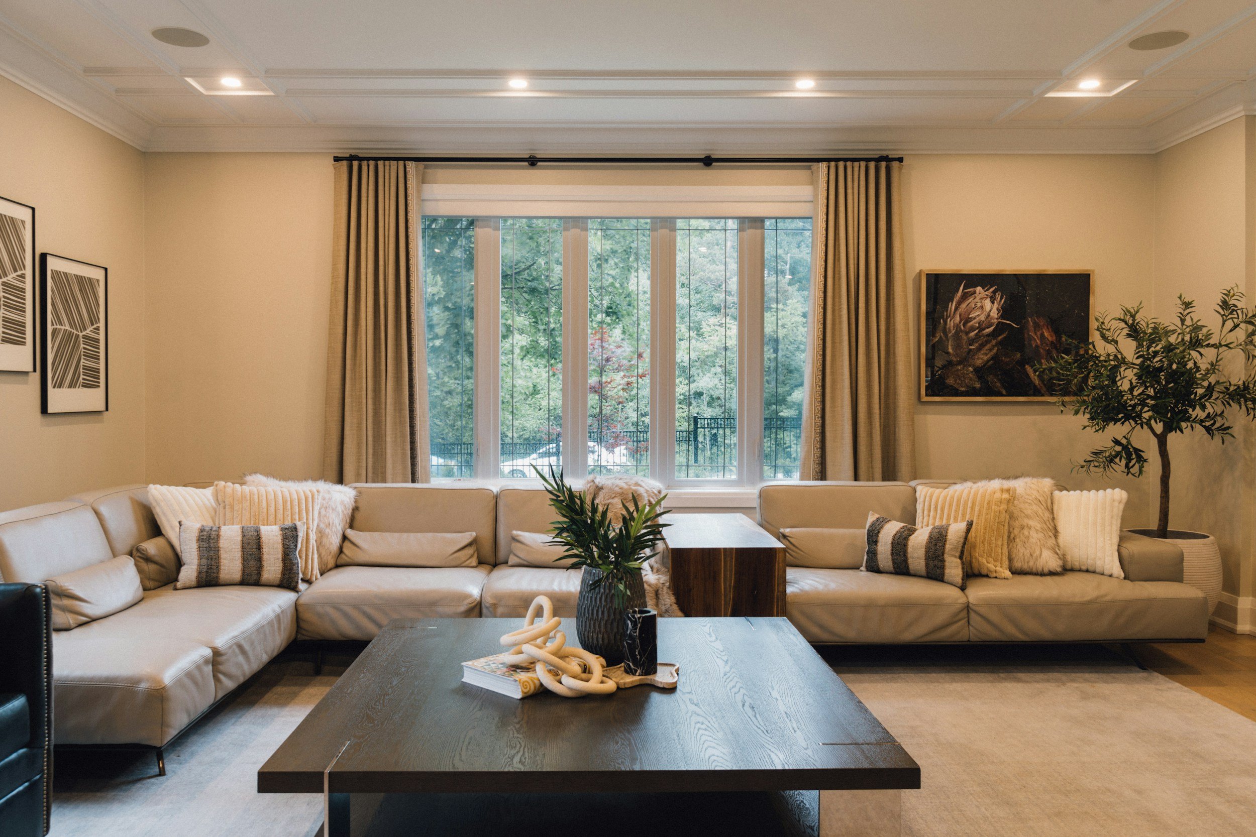 Living room with beige sectional sofa, pillows, a dark wood coffee table, a plant, wall art, curtains, and a large window showing greenery outside.