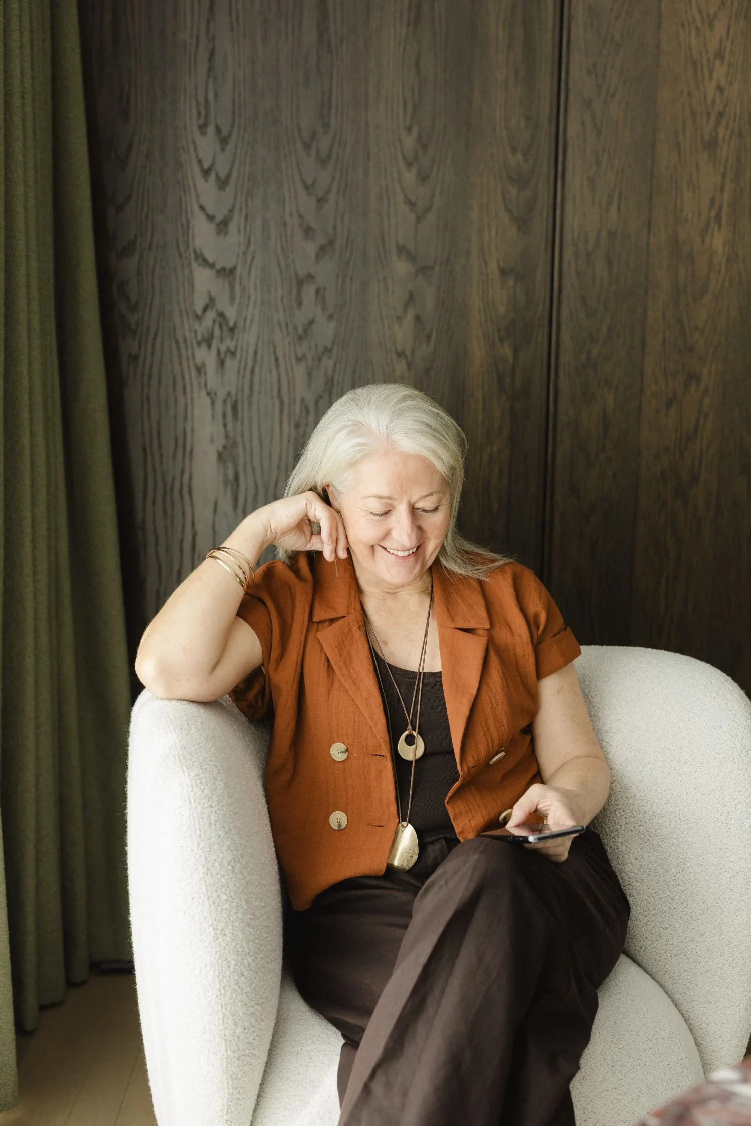 An older woman with gray hair, smiling and looking at her phone, seated in a white armchair, wearing a brown shirt with large buttons, gold jewelry, and a pendant necklace, in a room with a wooden wall and green curtains.
