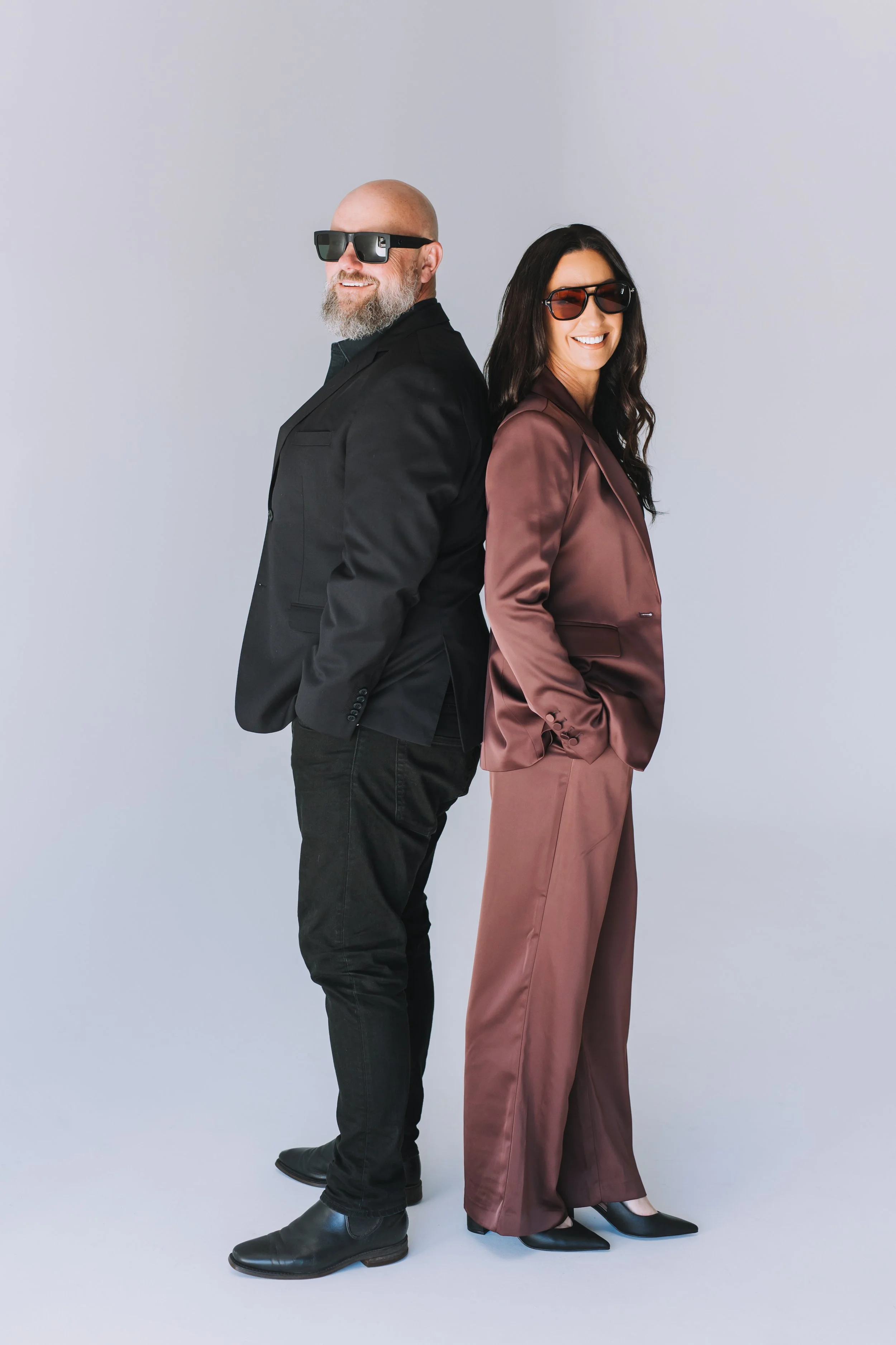 A man and woman standing back-to-back, smiling, wearing sunglasses in a studio with a plain background.