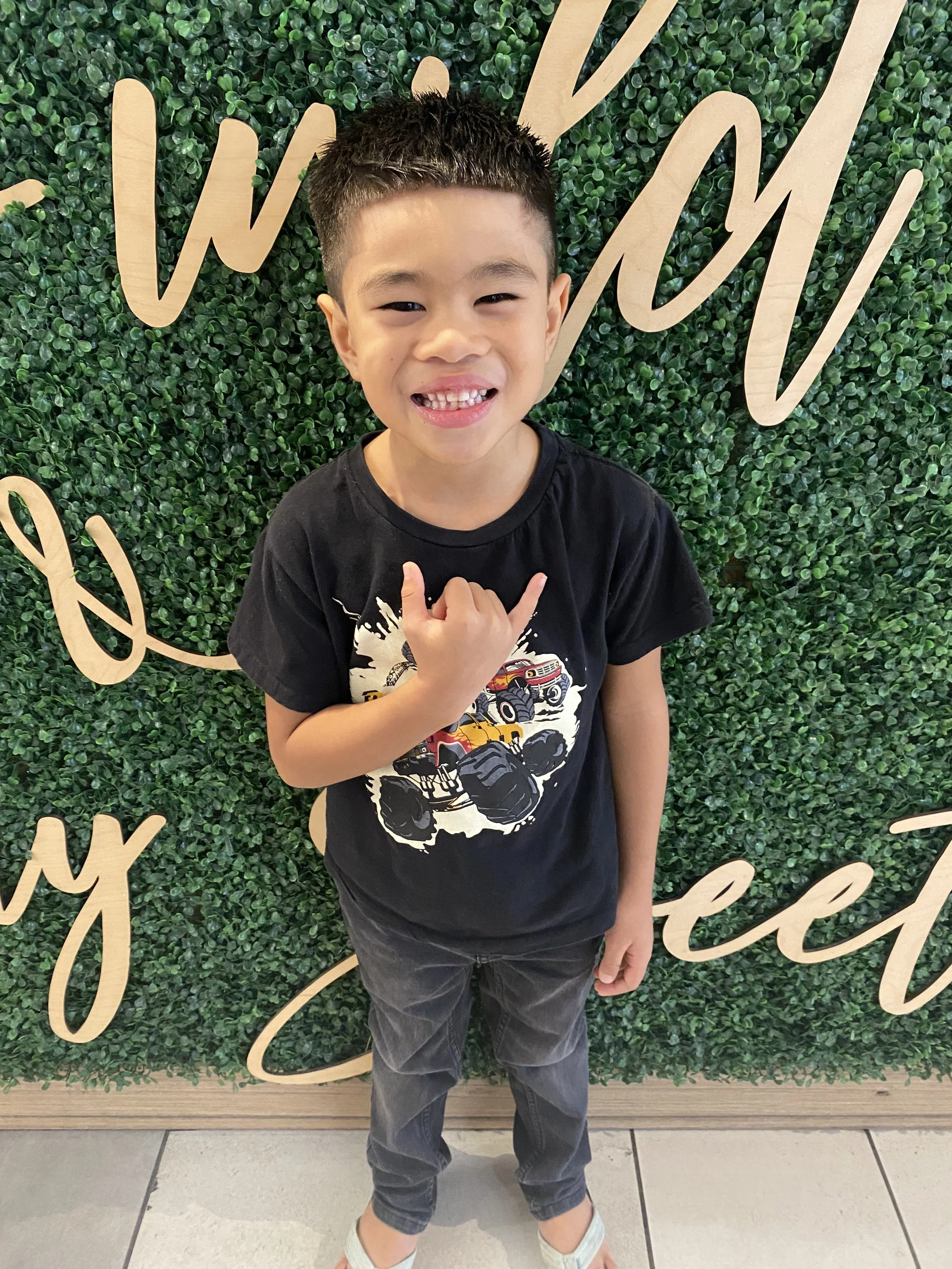 Young boy standing in front of a green leafy background with decorative writing, smiling and showing a hand gesture.