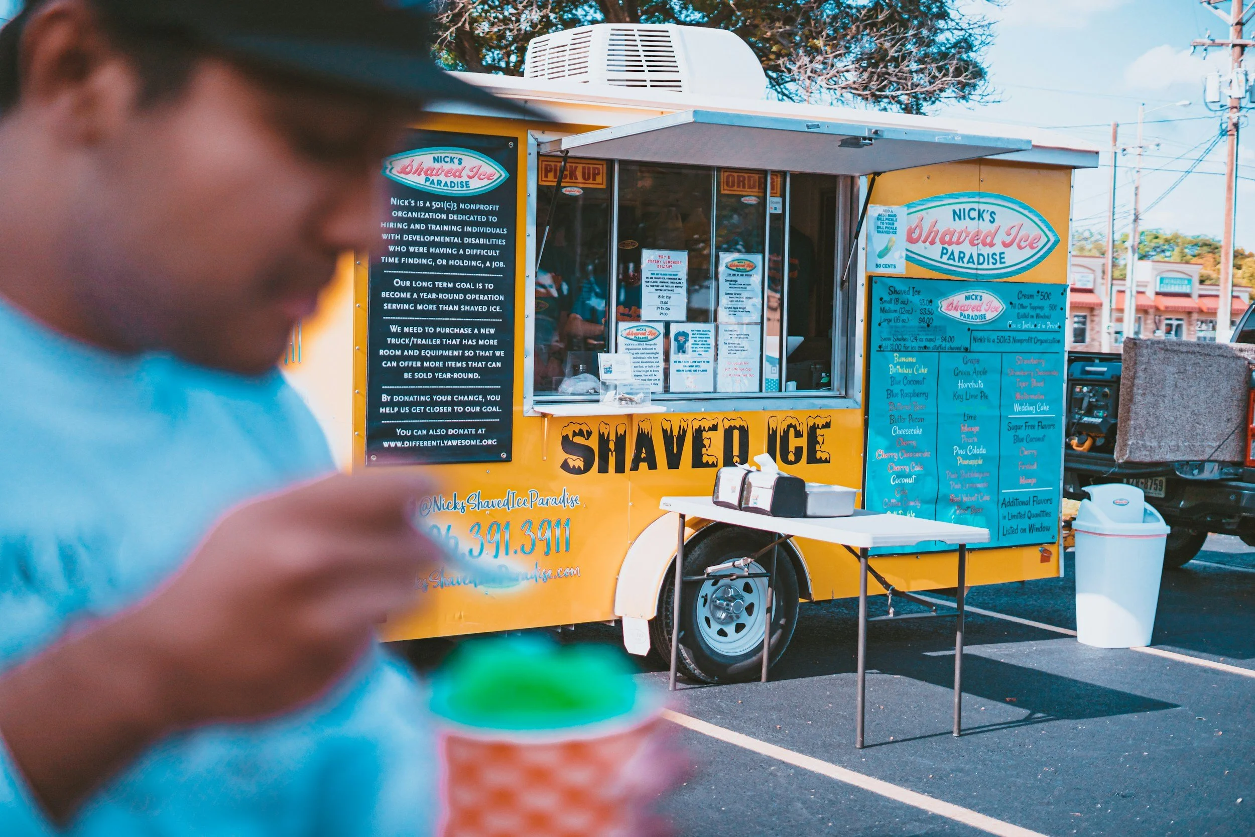 A person in the foreground with glasses and short hair, out of focus, with a yellow ice cream truck in the background. The truck has colorful signs and menus advertising shaved ice and other treats. There's a small table with pink napkins and a cart nearby, parked in a lot with utility poles and trees in the distance.