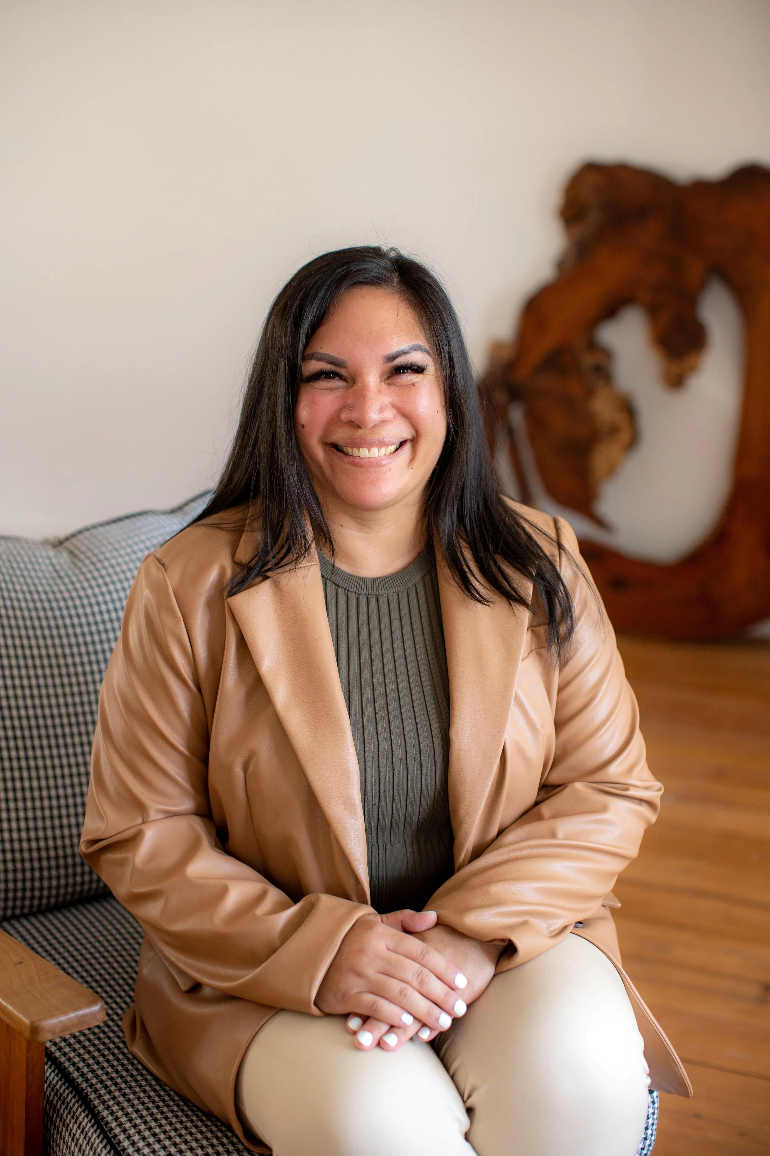 A woman with long dark hair and a big smile sitting on a checkered cushioned chair with a wooden armrest, against a plain wall with a large abstract wooden sculpture in the background.