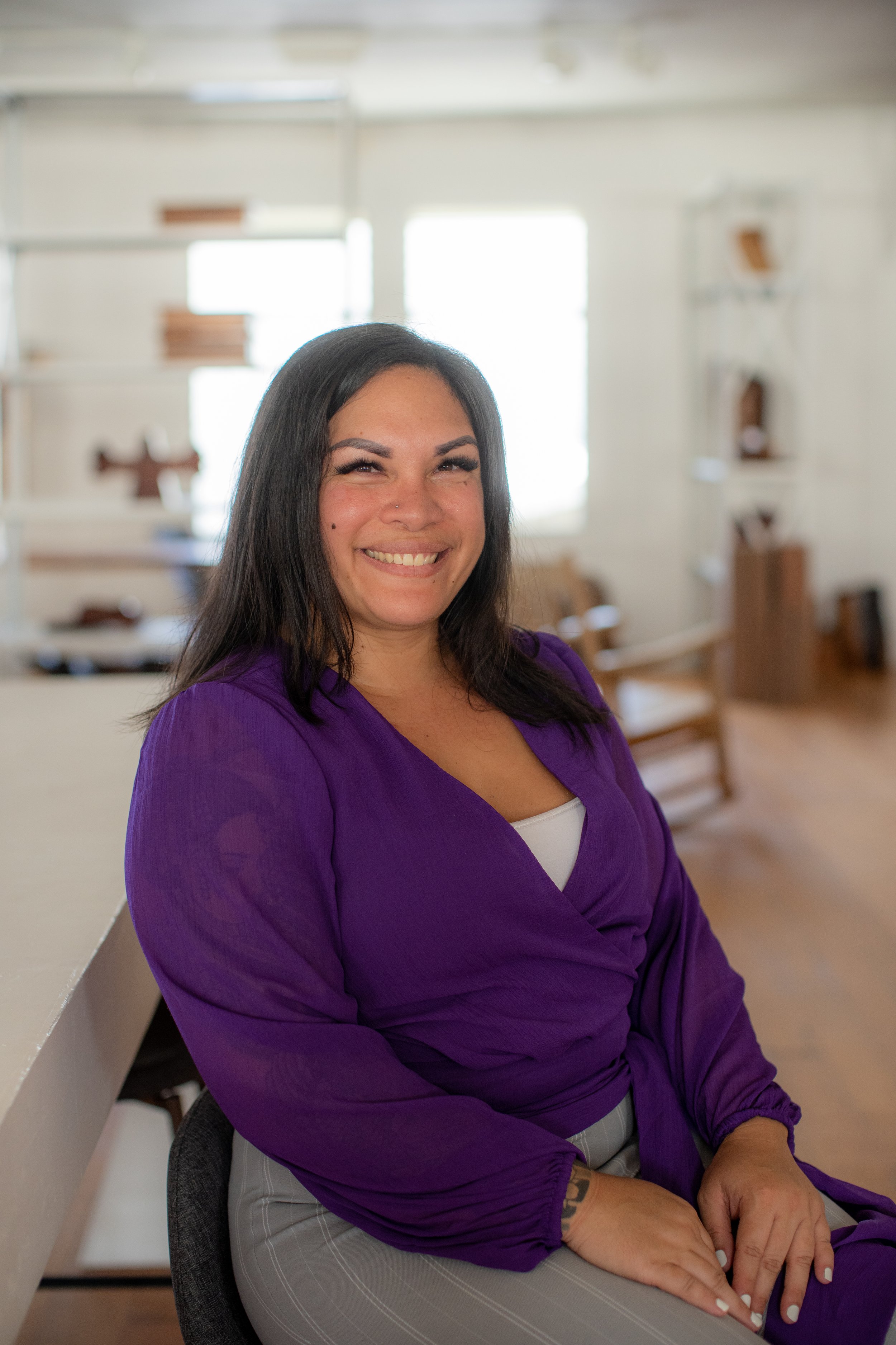 A woman with dark hair smiling, wearing a purple blazer over a white top, sitting in a well-lit room with large windows and white shelves in the background.
