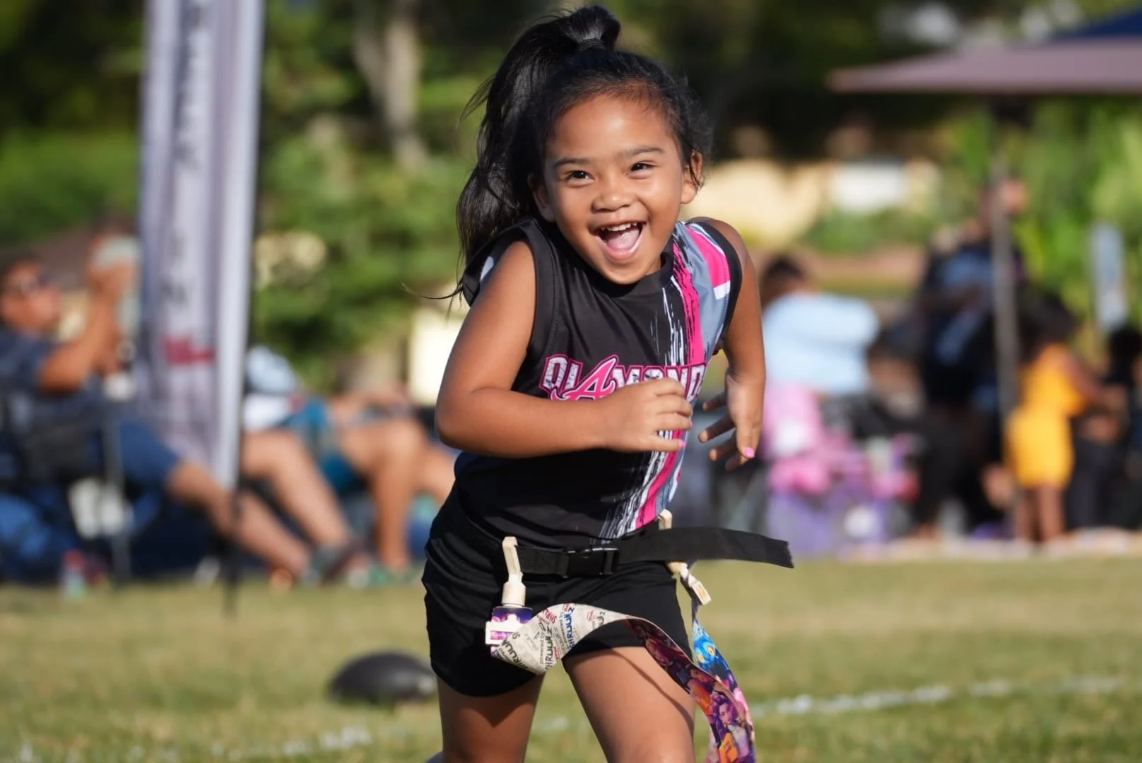 A young girl with a ponytail running outdoors on grass during daytime, smiling cheerfully, with people and trees blurred in the background.