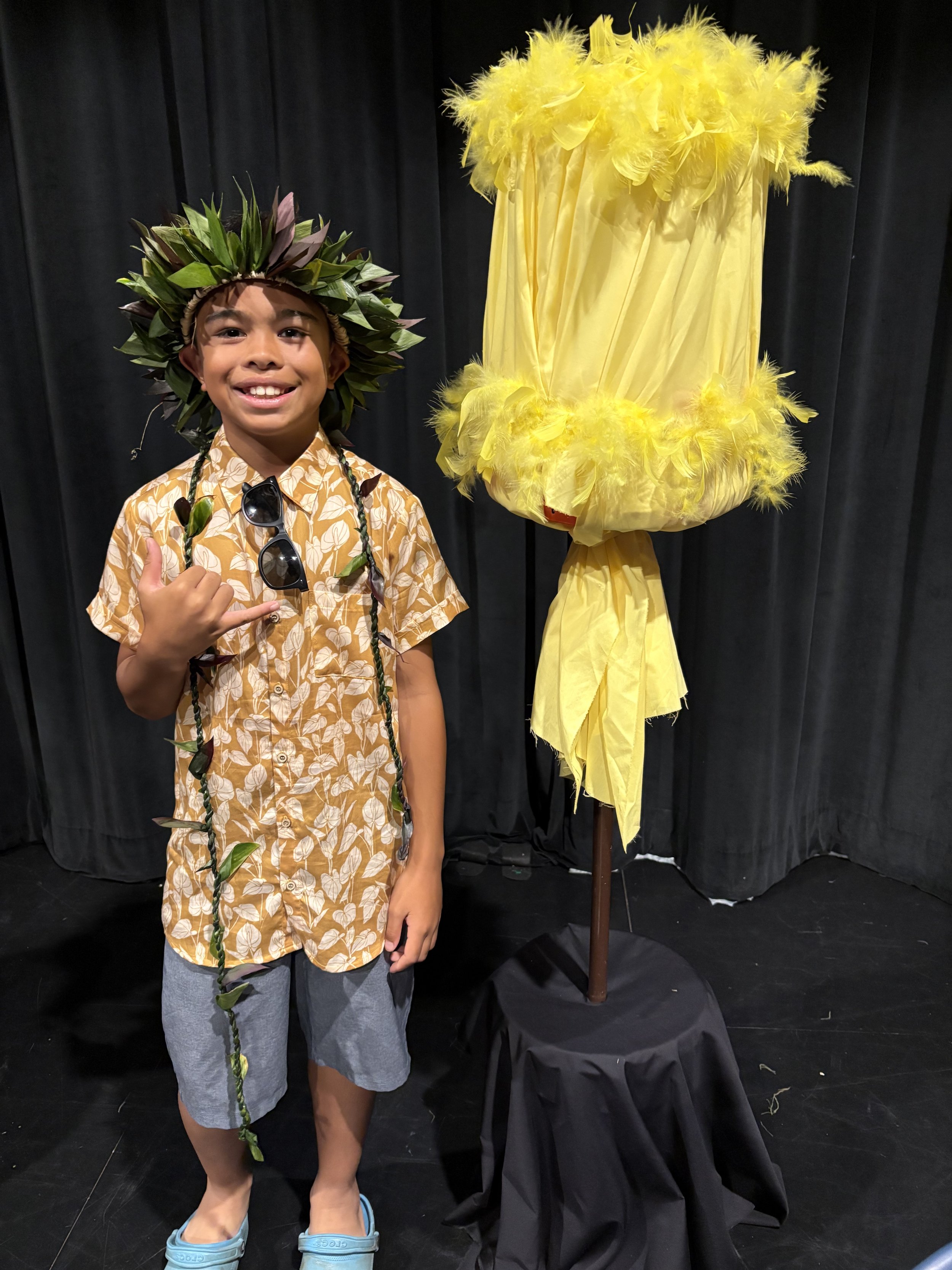 A young boy smiling and giving a thumbs-up, wearing a leaf crown and a Hawaiian shirt, standing next to a yellow decorative fabric on a stand against a black curtain background.