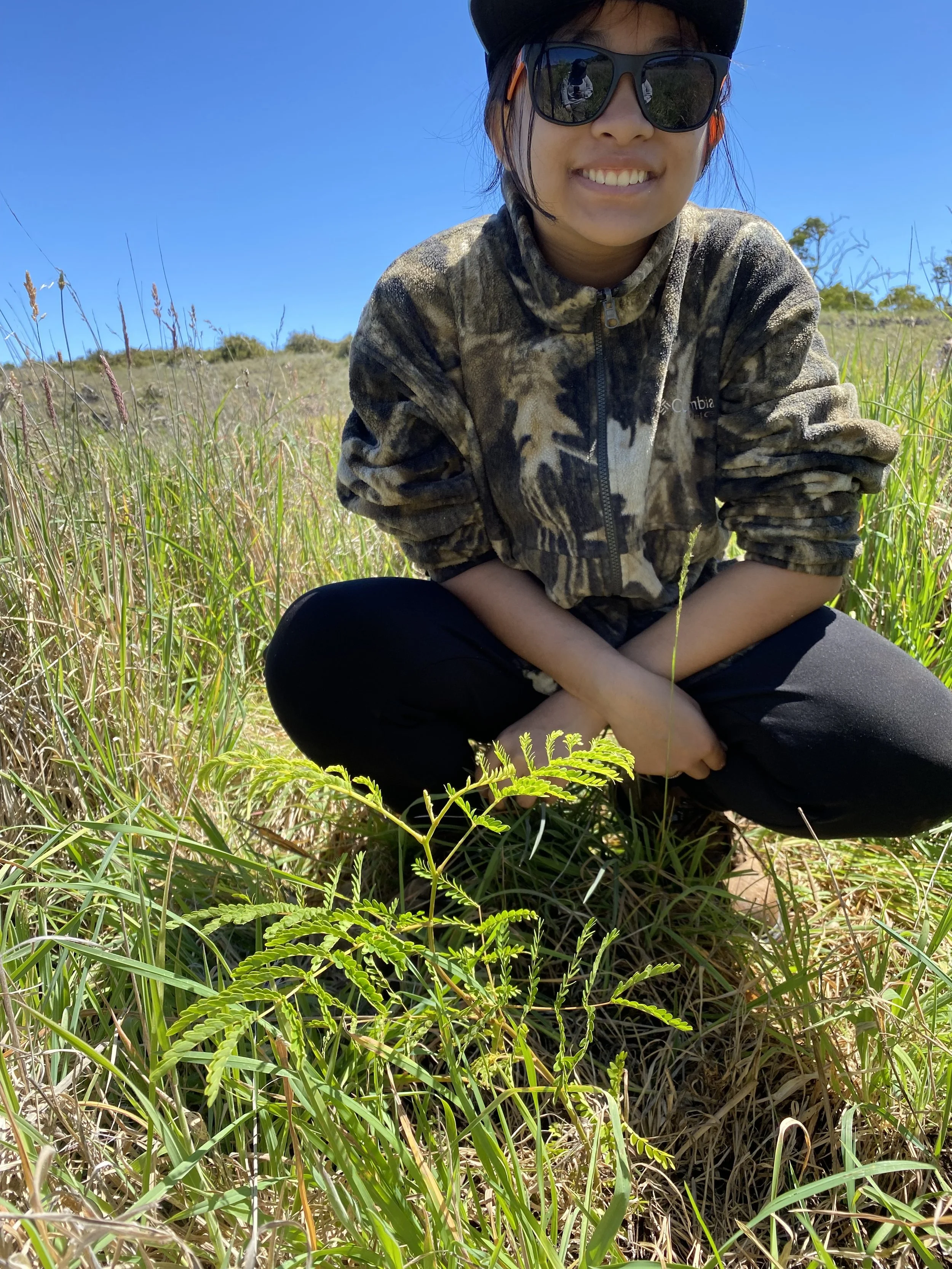 A woman smiling, wearing sunglasses, a camouflage jacket, and a hat, sitting in a grassy outdoor area with clear blue sky and some bushes in the background.
