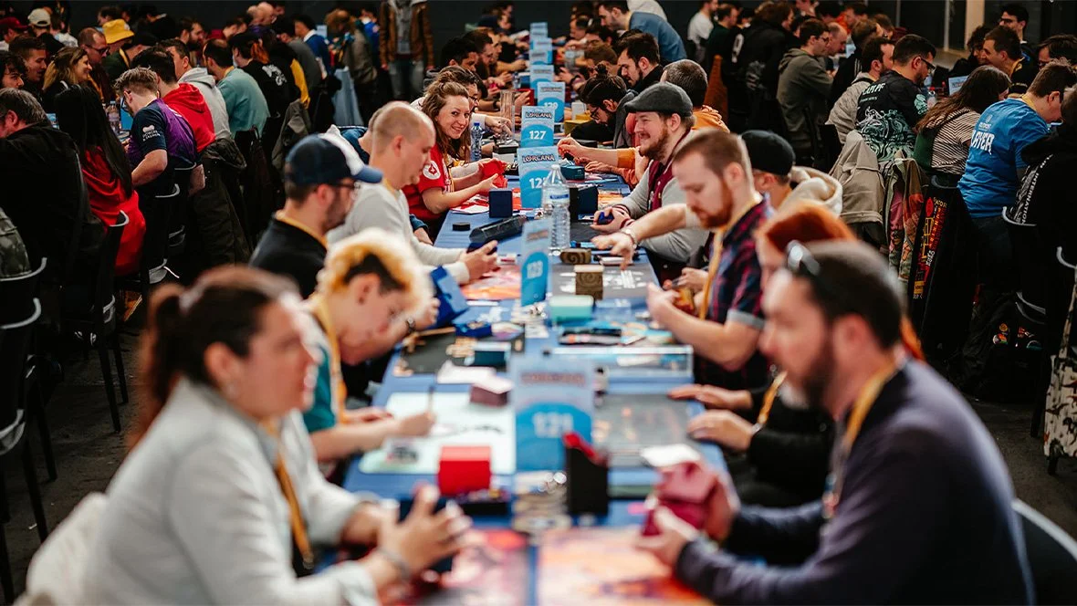 People playing a card game at a large table during a gaming tournament or convention.