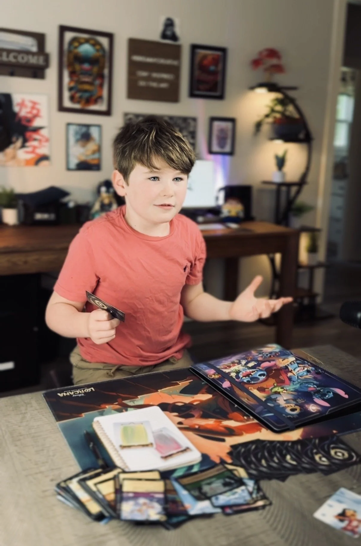 A young boy with brown hair wearing a red t-shirt, sitting at a table with Disney Lorcana and a tablet, in a room decorated with framed artwork and shelves with plants and decorations.
