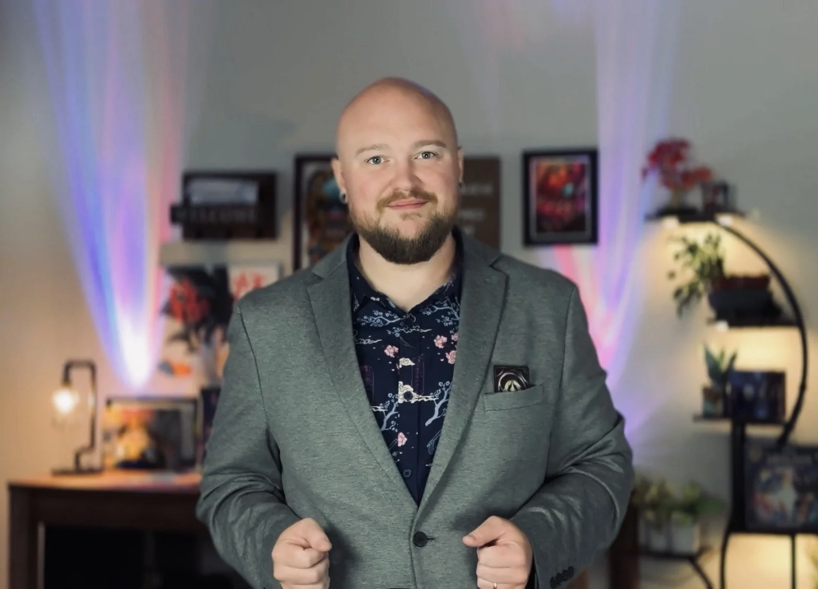 A man in a gray blazer and patterned shirt is smiling at the camera, standing in a decorated room with shelves, framed pictures, and colorful LED lights in the background.