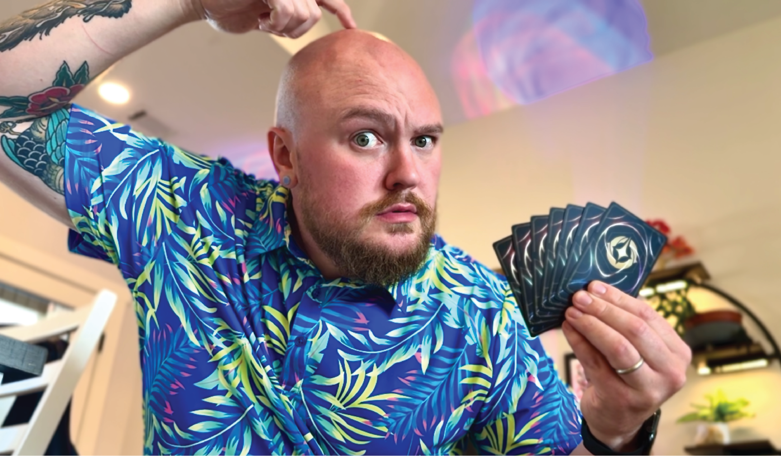 A man with a beard, bald head, and tattoos on his right arm wears a colorful, patterned shirt and holds a set of tarot cards while looking at the camera