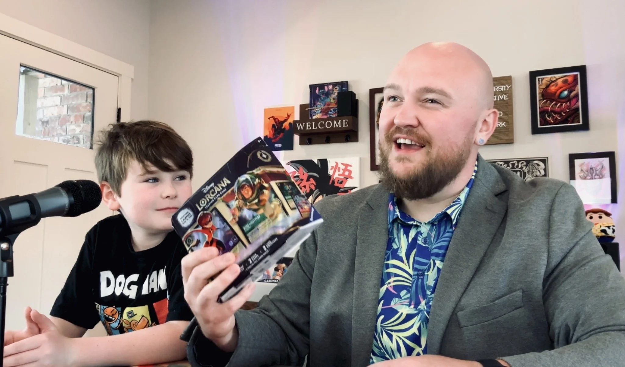 A man with a beard and bald head smiling as he receives a toy from a young boy with brown hair. The man is holding a box of Disney Lorcana's 2-Player Starter Set featuring "The Incredibles" and "Toy Story". They are indoors with framed artwork.