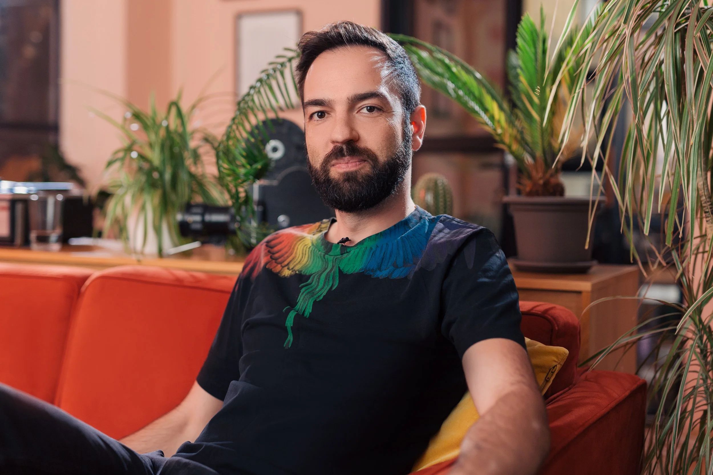 A young man with dark hair and a beard sitting on an orange couch surrounded by large green plants in a cozy indoor setting.