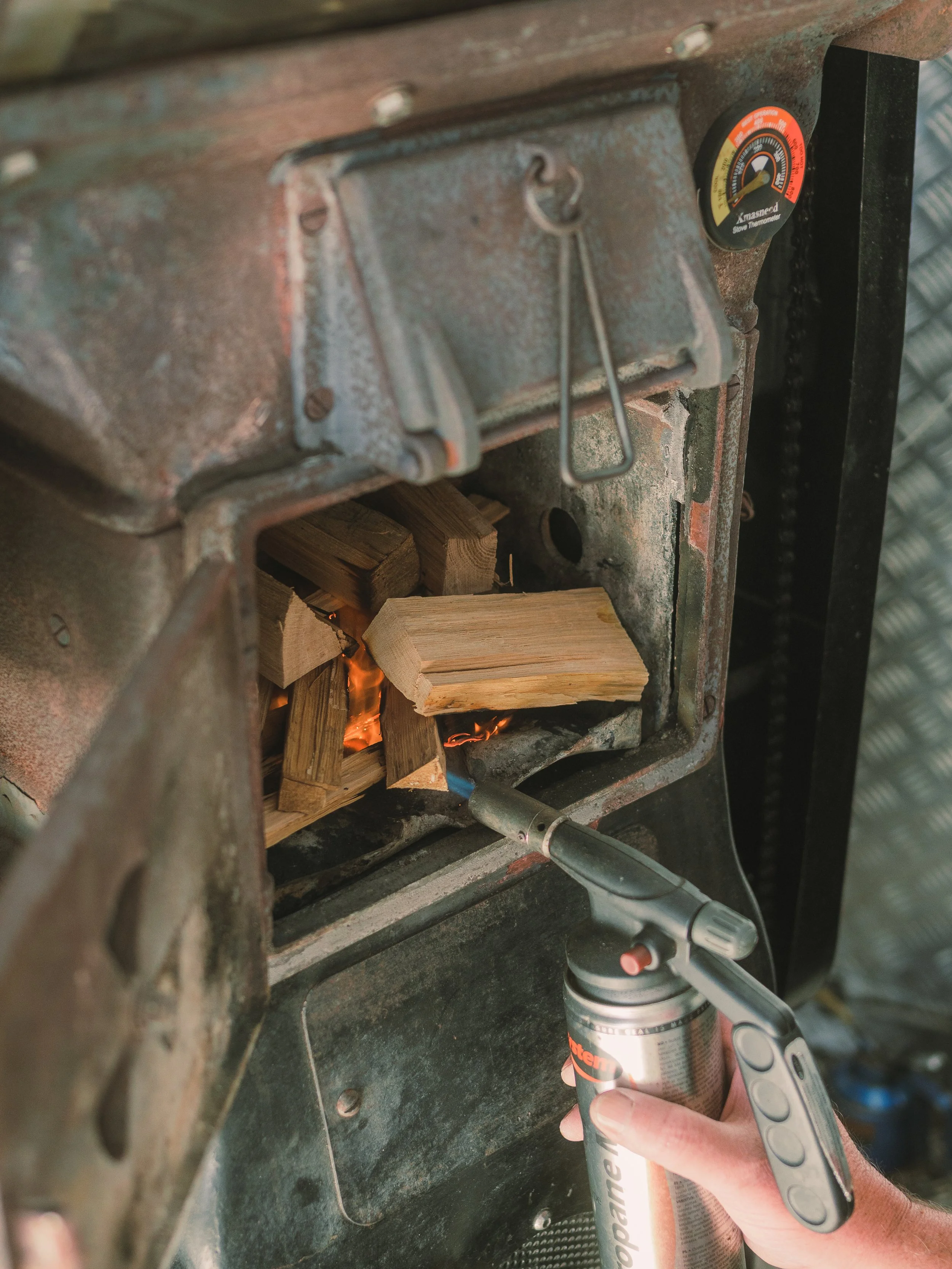 Person using a blowtorch to ignite wood in a firepit with a metal structure, wood pieces, a thermometer gauge, and a propane tank.