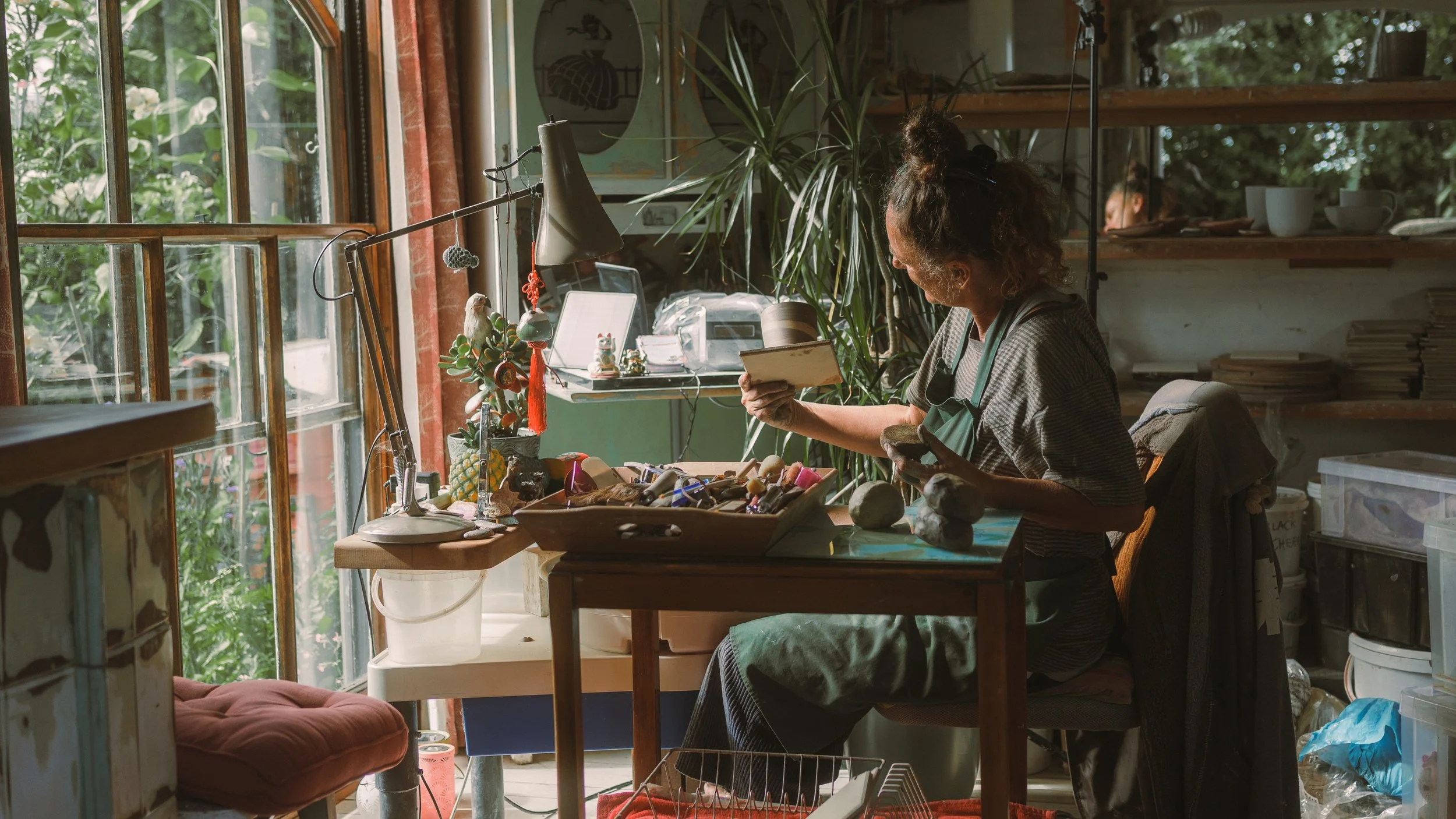 A woman with curly hair and a bun, wearing an apron, working at a cluttered craft table in front of a large window with trees and plants outside. The table has various art supplies, rocks, and decorative items, with a large plant behind her.