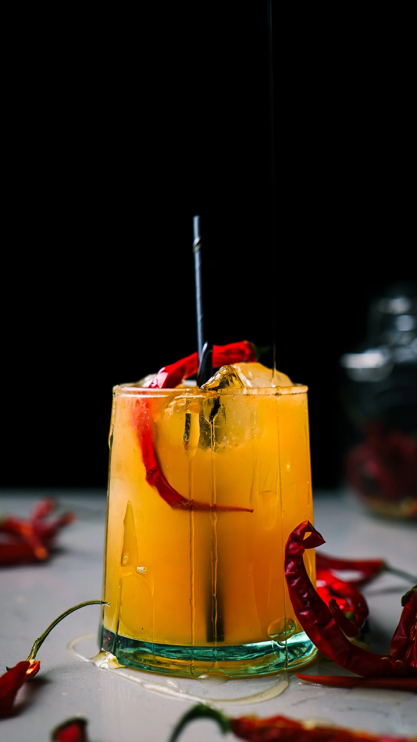 A tall glass of orange-colored cocktail with ice cubes, garnished with red chili peppers and a cherry, on a white surface with spilled liquid and chili peppers around.