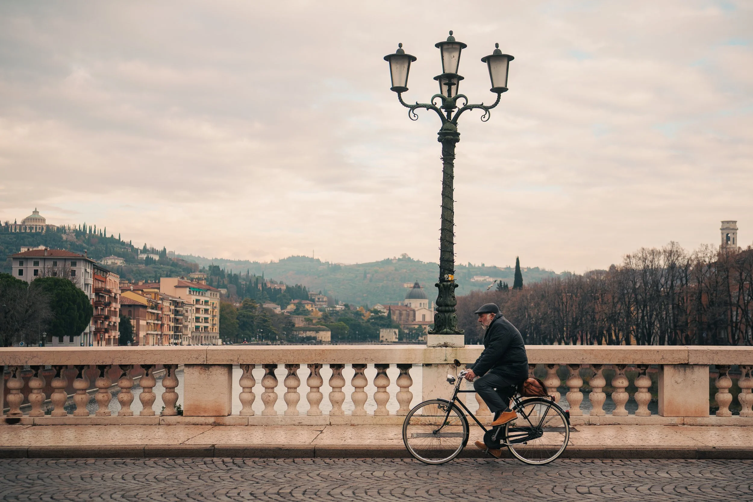 A man riding a bicycle along a stone bridge with a railing, under a streetlamp with four lanterns. In the background, there are residential buildings, hills with trees, and a cloudy sky.