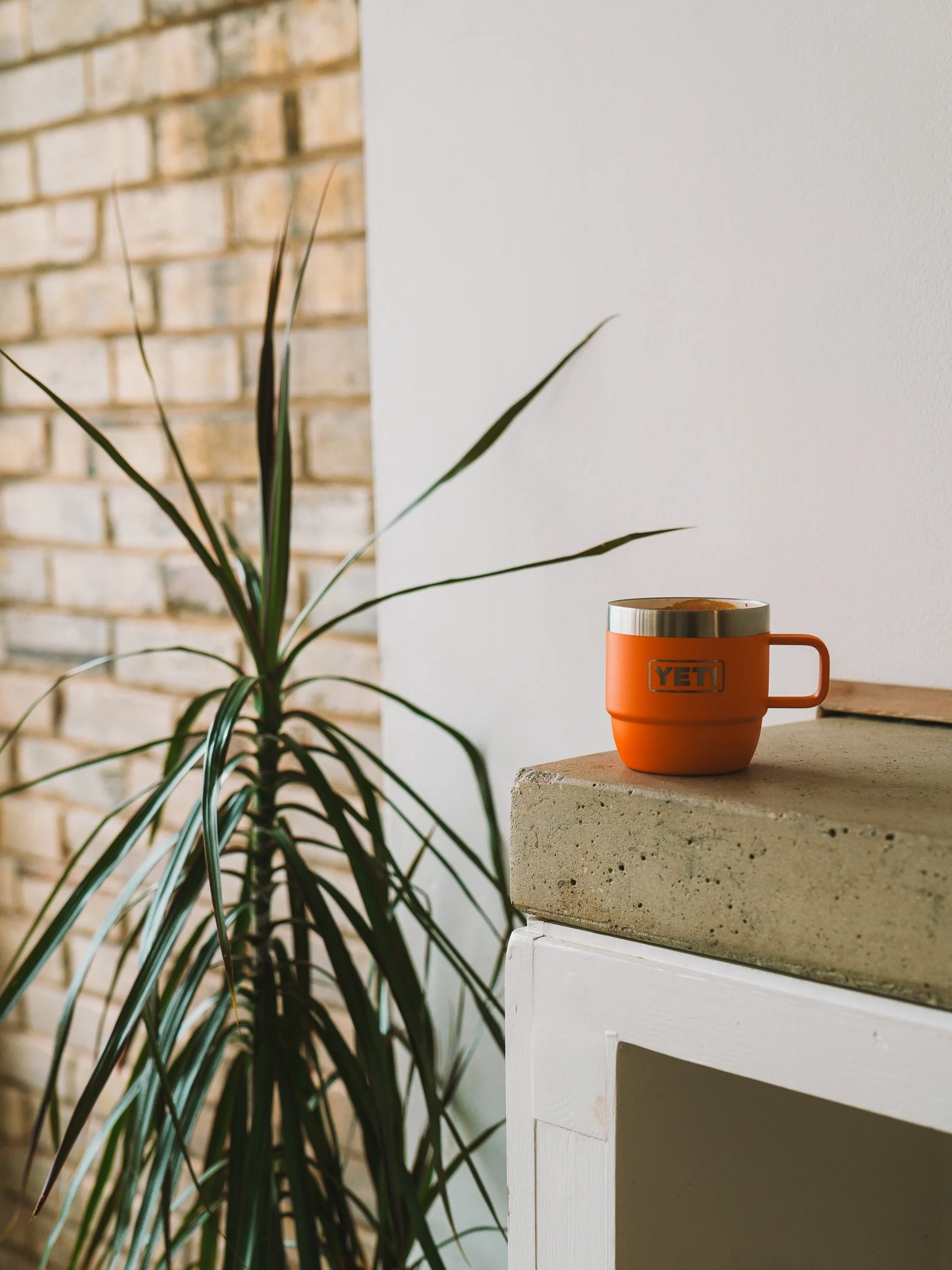 An orange YETI mug on a light brown wooden surface with a white cabinet beneath, positioned beside a green leafy houseplant in front of a brick wall and white wall background.