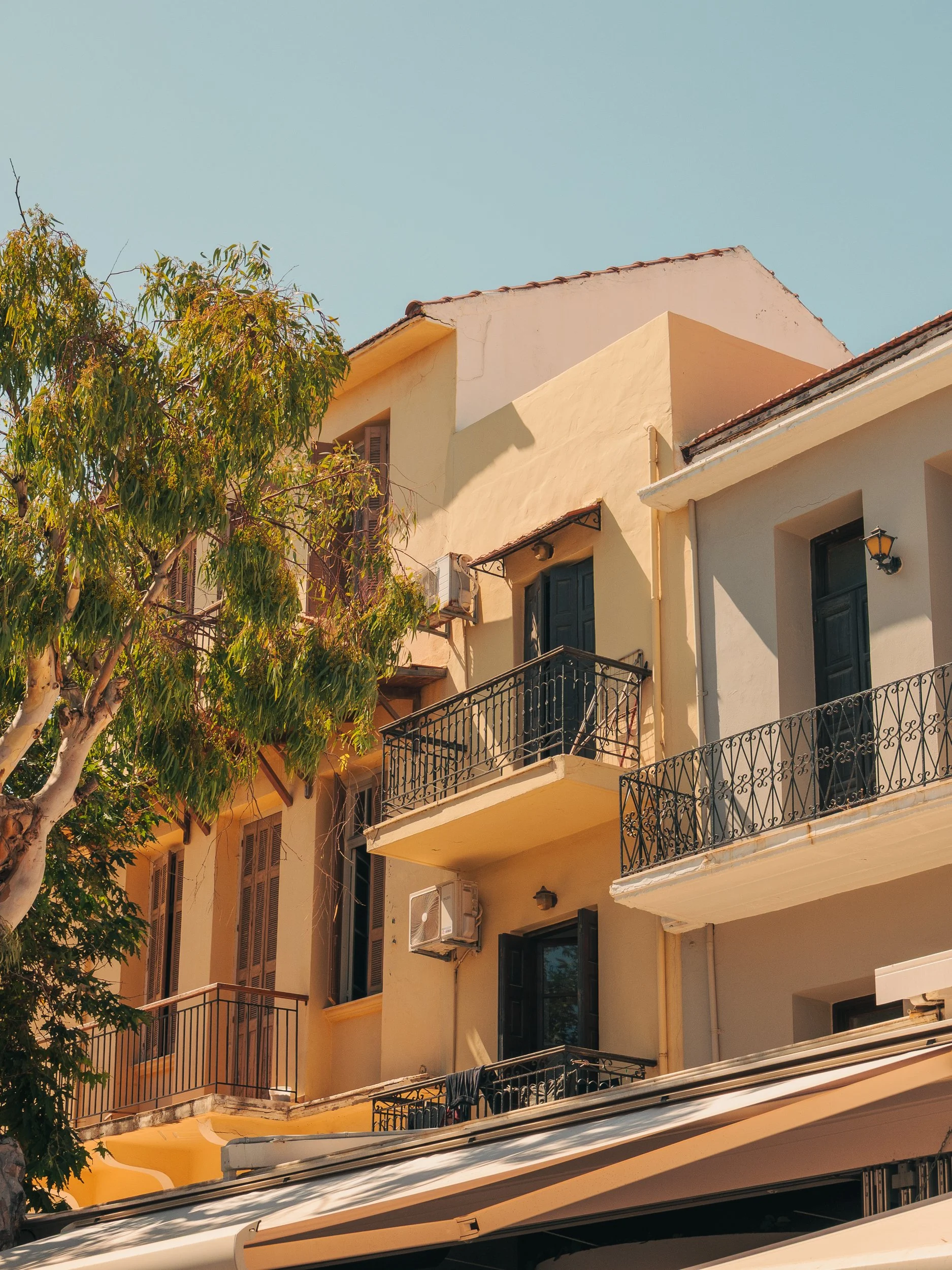 Multi-story yellow building with black balconies, windows, and air conditioning units, partially shaded by a large tree, under a clear blue sky.