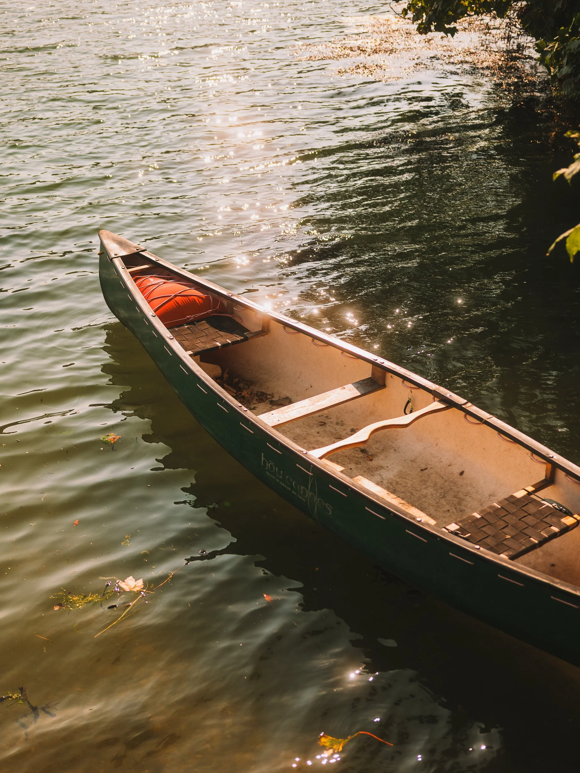 Partially submerged small boat in a lake or river with water reflecting sunlight, some floating leaves, and a shoreline with greenery on the right side.