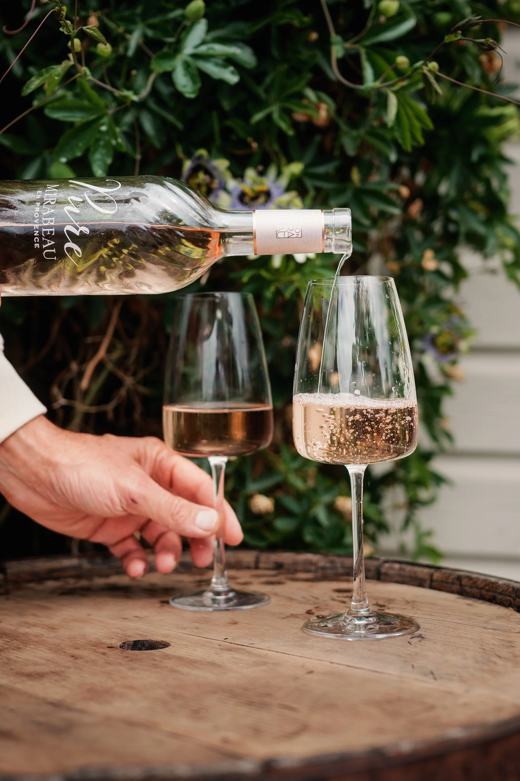 A person is pouring rosé wine from a bottle into two wine glasses on a wooden table outdoors, with a background of green foliage and purple flowers.